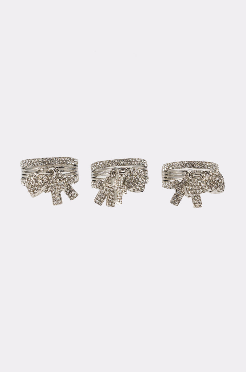 The ANGELS WITH ME RING SET features three silver rings adorned with small gemstones and charm pendants shaped like ribbons and hearts, all displayed on a plain white background—a charming rhinestone jewelry collection. 1