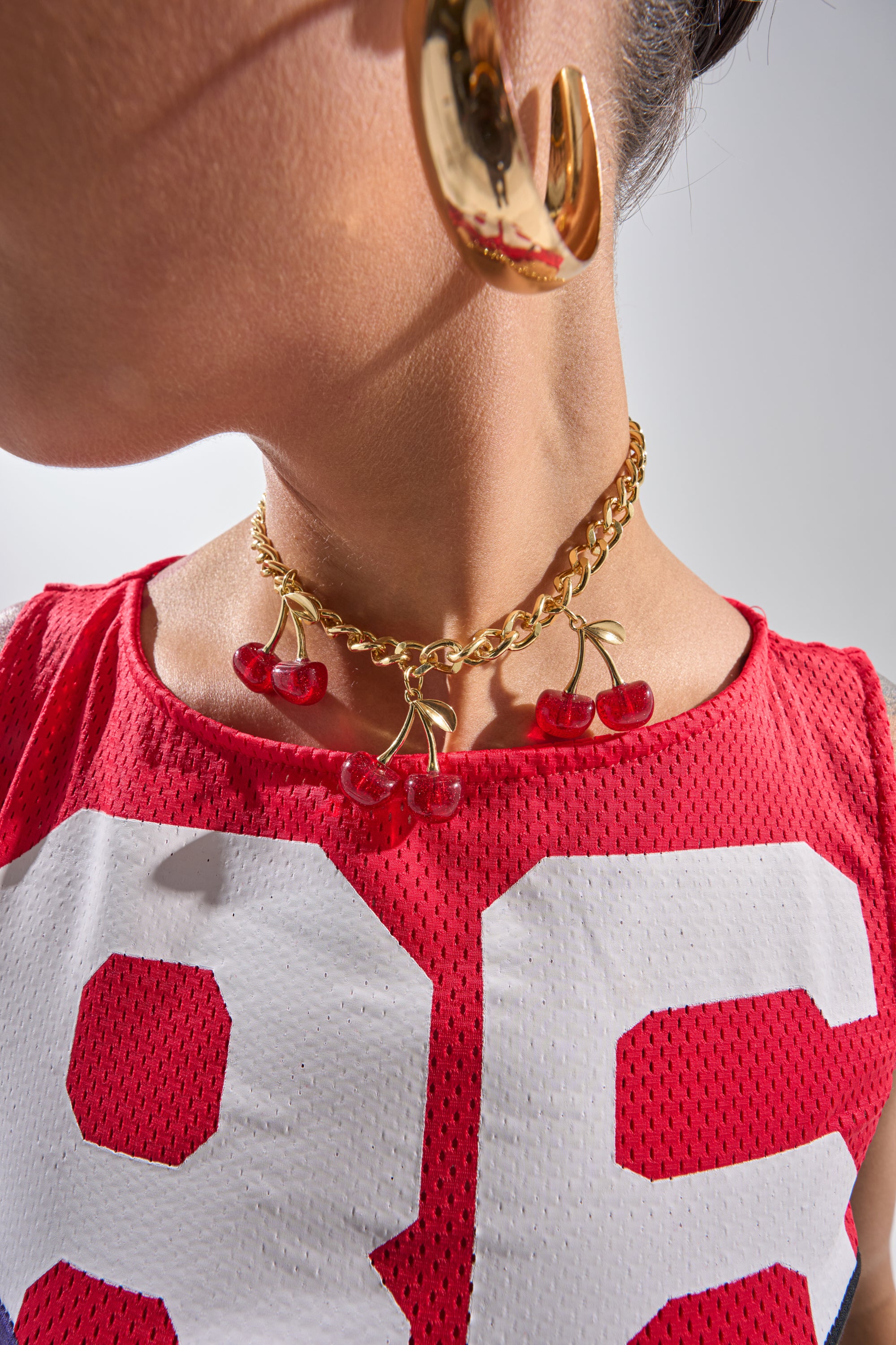 A close-up of a person wearing the POP IT NECKLACE, a Y2K-inspired gold cherry pendant, paired with large gold hoops and a red mesh top featuring the number 85 in white. Their face is partially out of frame. 0