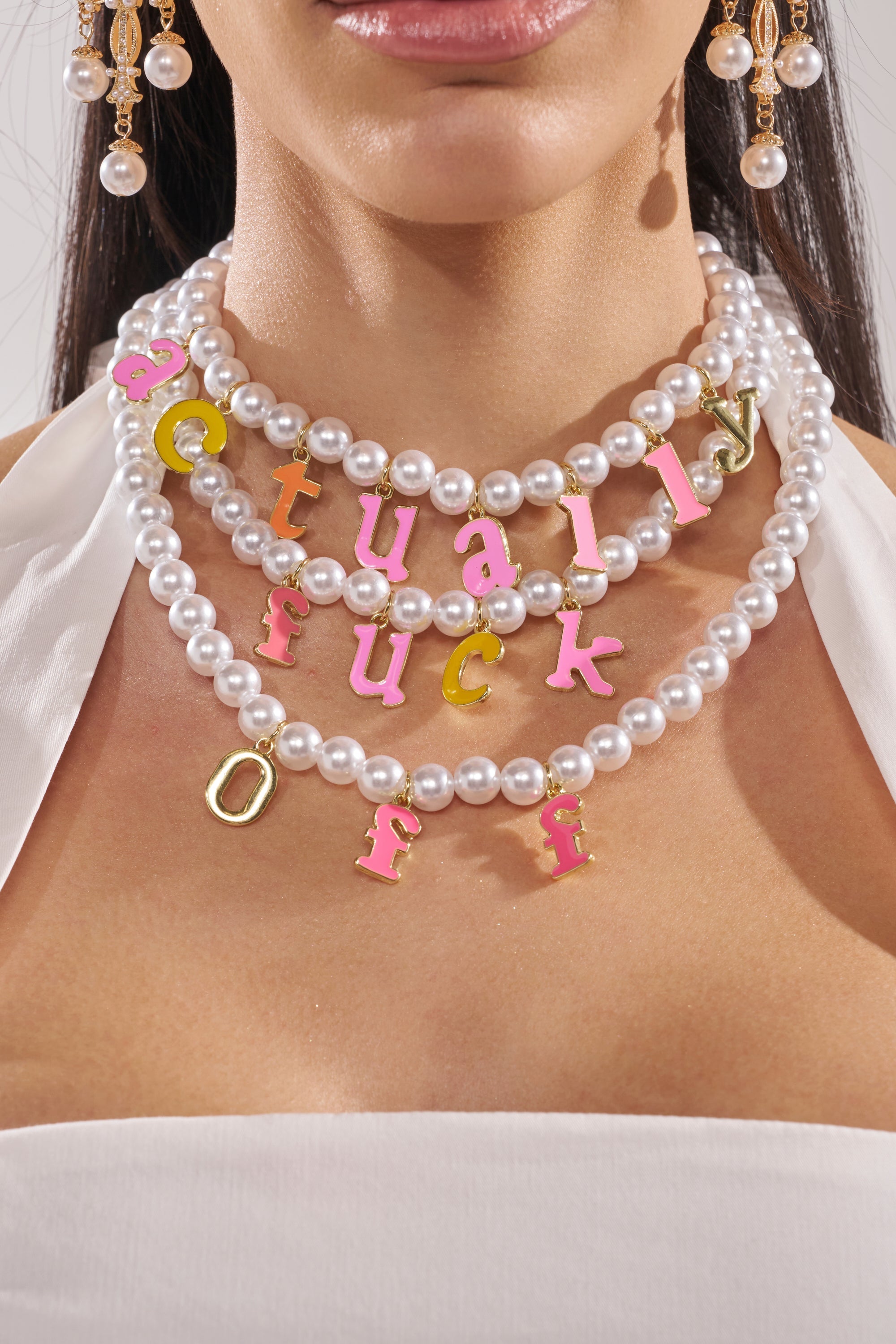 A woman wears a white halter top and the I SAID WHAT I SAID NECKLACE, featuring faux pearls and colorful enamel charms spelling "actually fuck off." She finishes the bold look with gold drop earrings accented with pearls. 1