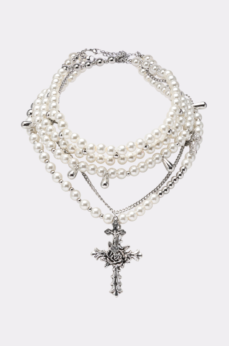 The LAYERED UP CROSS NECKLACE features layers of white pearls, silver chains, and cross charms, highlighted by a prominent ornate cross pendant with a rose detail, shown against a plain light background. 1