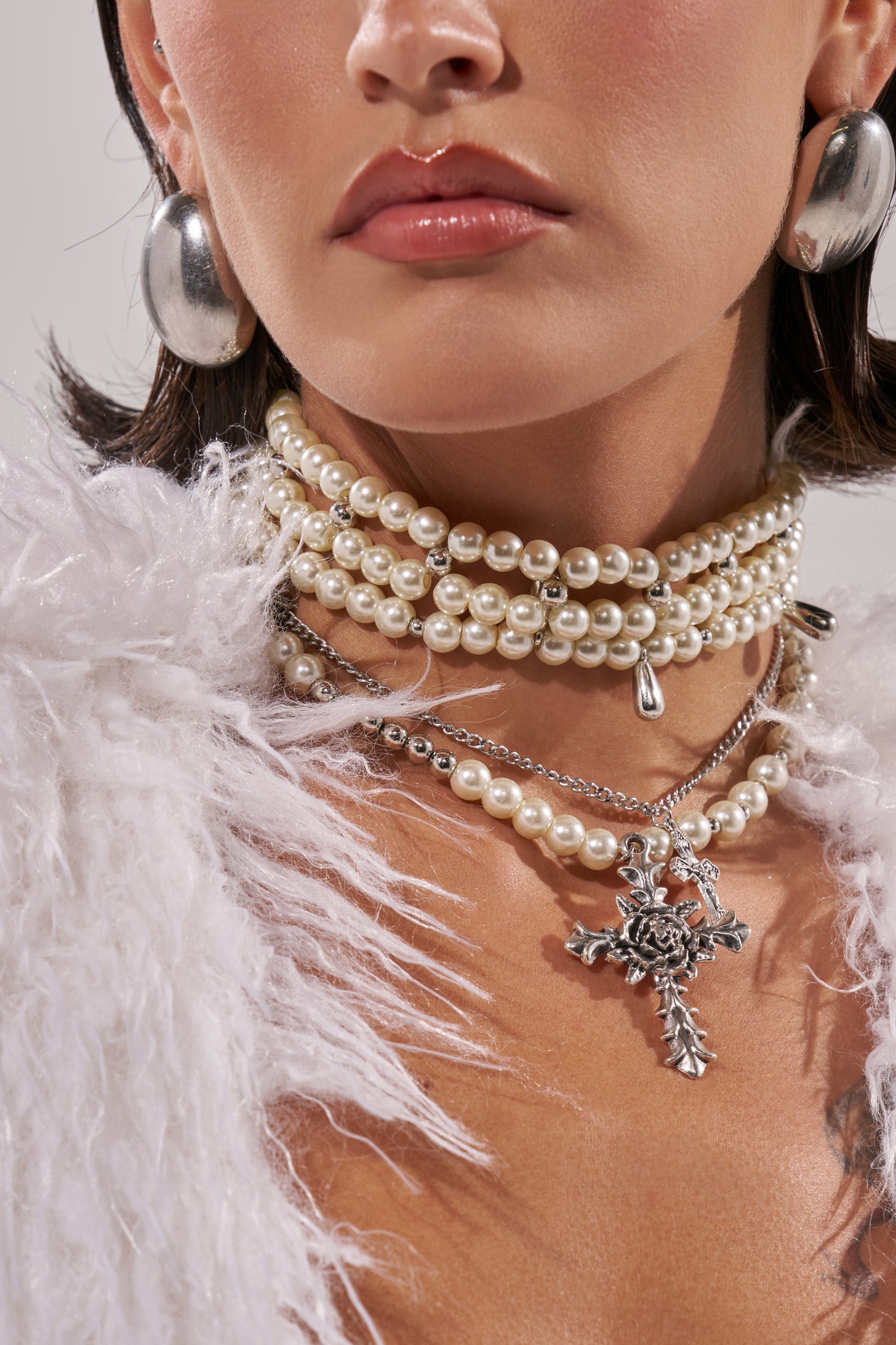 A woman wears the LAYERED UP CROSS NECKLACE, featuring layered pearls and a bold silver cross pendant, along with gothic jewelry and large silver earrings. She dons a white fluffy-textured garment; the photo centers on her neck and jawline. 0