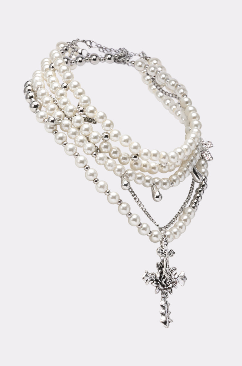 The LAYERED UP CROSS NECKLACE showcases five layers of faux pearl strands and silver chains with cross pendants and metallic accents, all arranged elegantly on a plain white background. 2