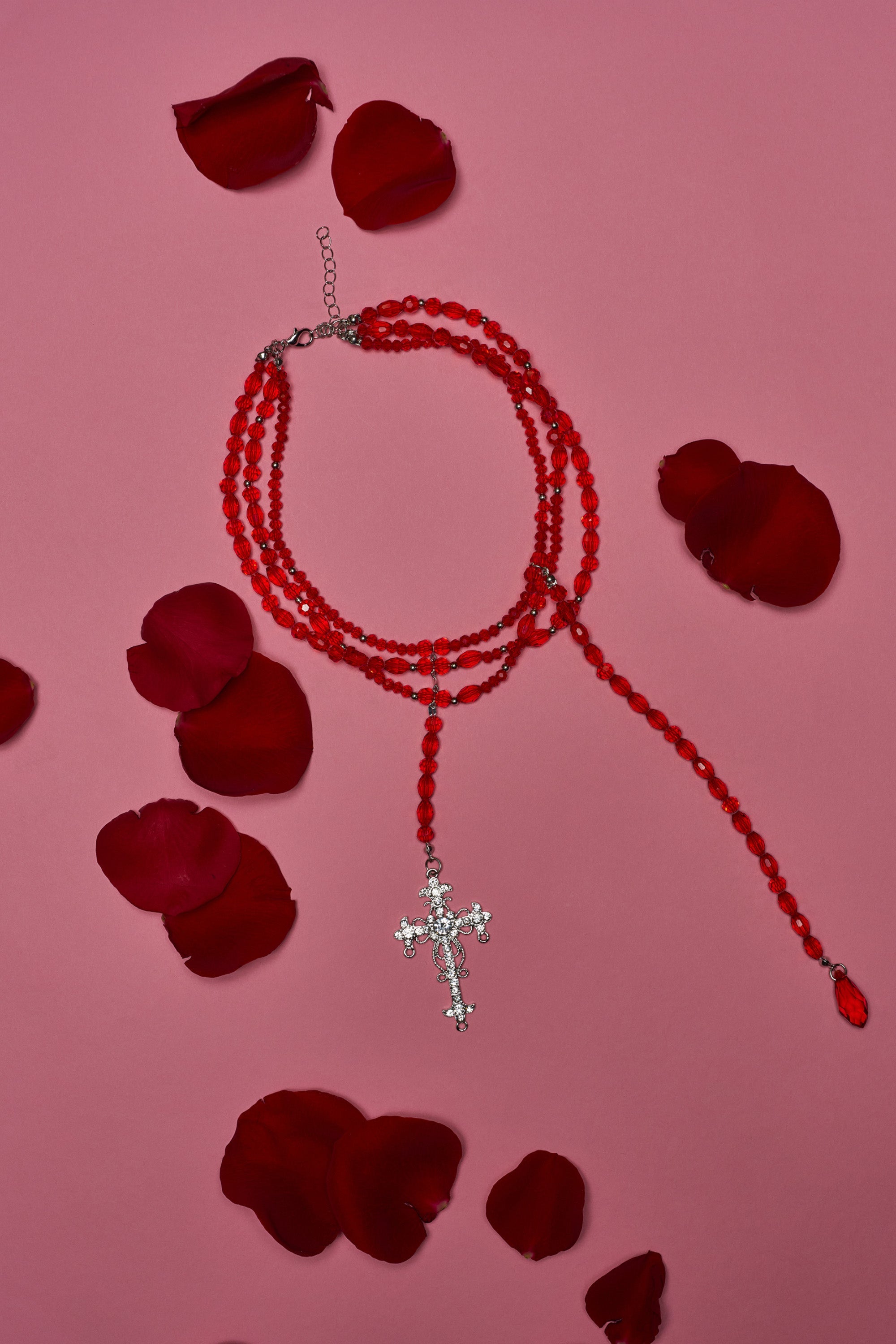 The TRIPPIE CROSS NECKLACE, a bold red beaded statement piece with a silver cross, is displayed in a circle on a pink background, surrounded by scattered dark red rose petals. 1