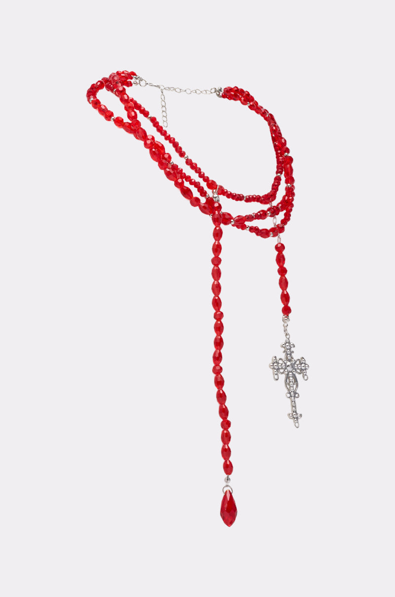 The TRIPPIE CROSS NECKLACE showcases layered red beads in three strands, finished with a silver ornate cross and a red teardrop bead, all presented on a plain white background. 4