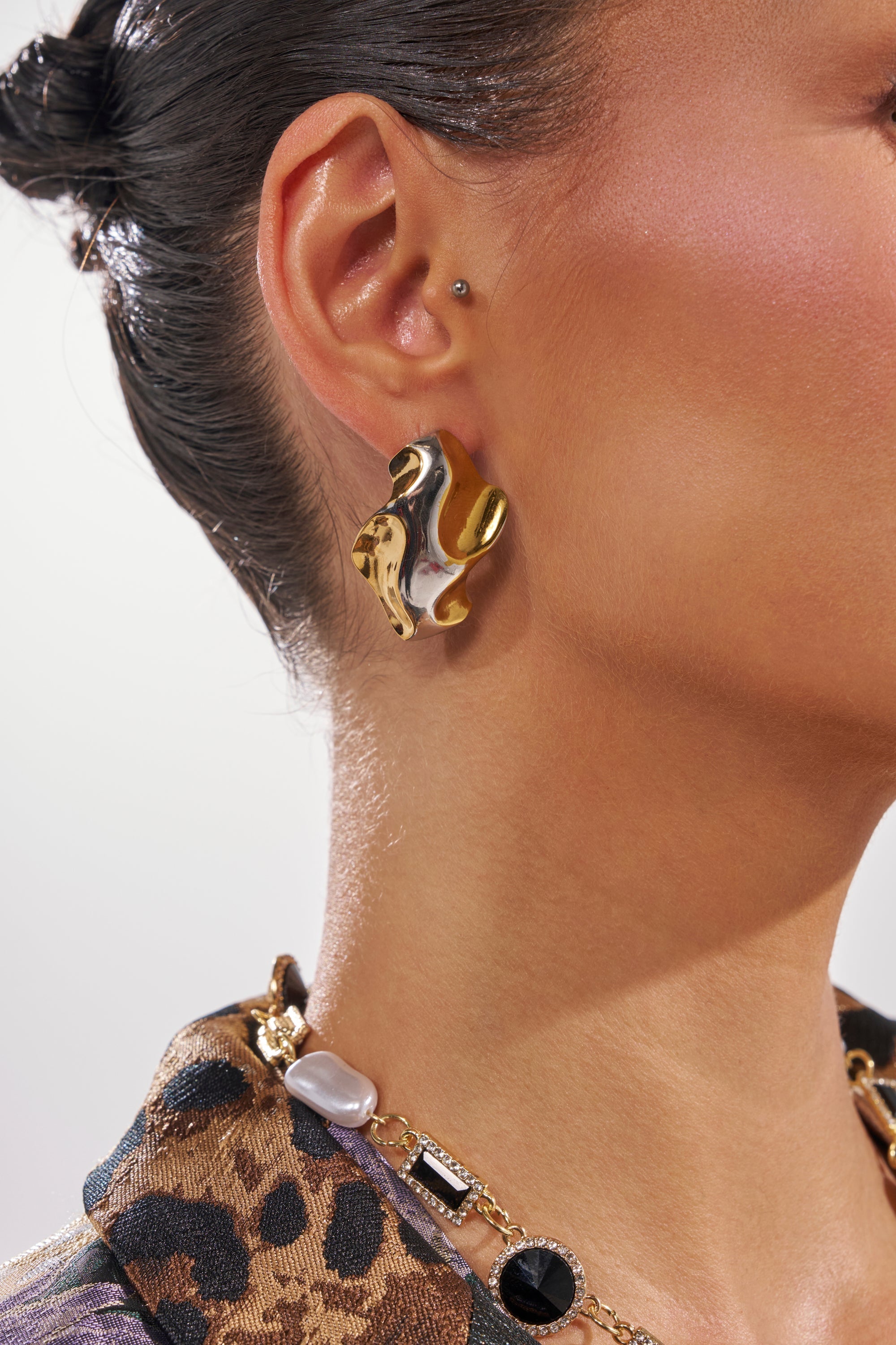 A close-up of a person wearing the MIXED FEELINGS EARRING—wavy, mixed-metal gold and silver—paired with a pendant gold chain necklace and a leopard-print top, hair pulled back. 1
