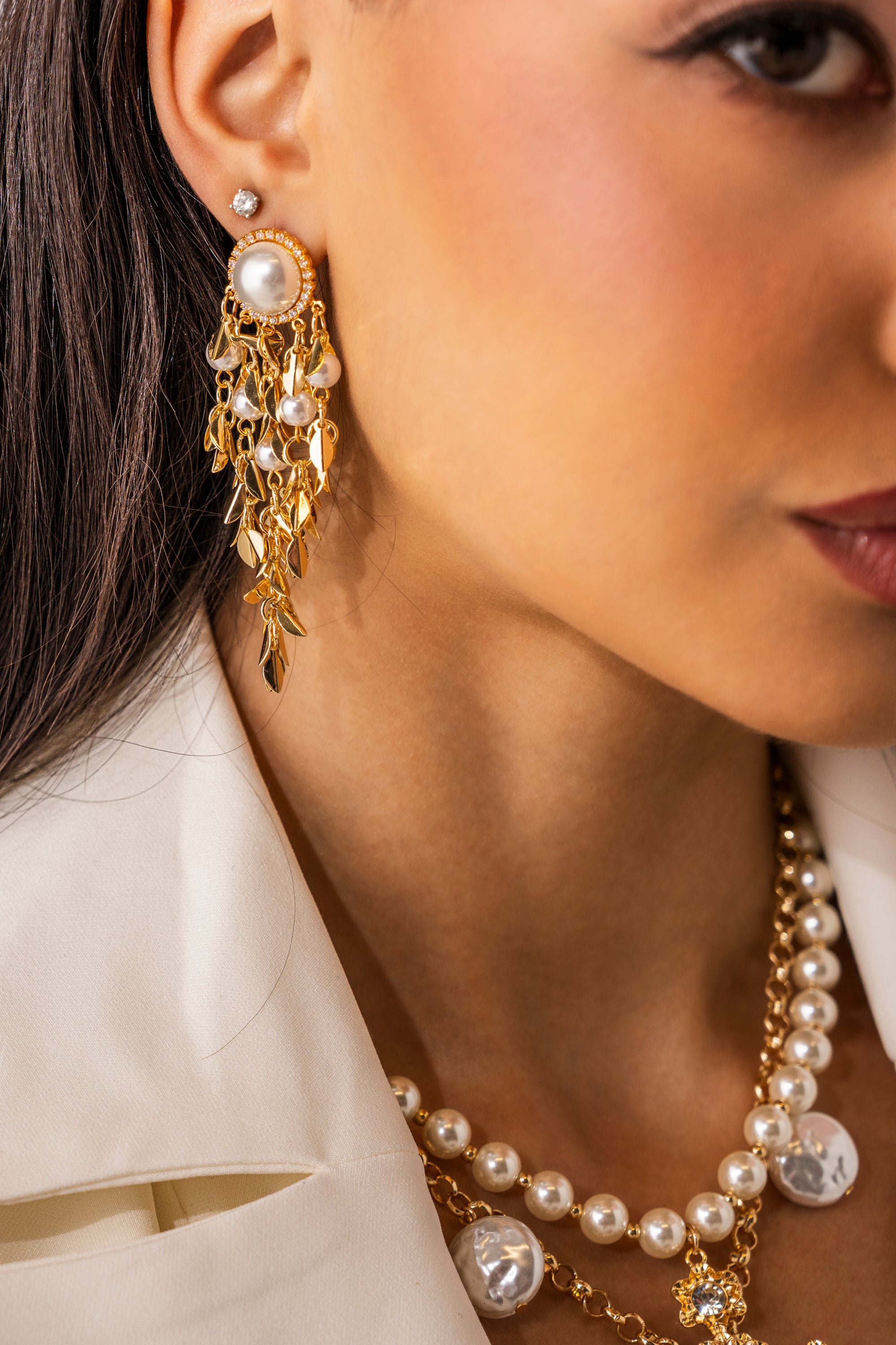 A close-up of a woman wearing the PARADISE EARRING—statement gold and pearl earrings—alongside a diamond stud, layered pearl and gold necklaces, all styled with a white blazer. 0