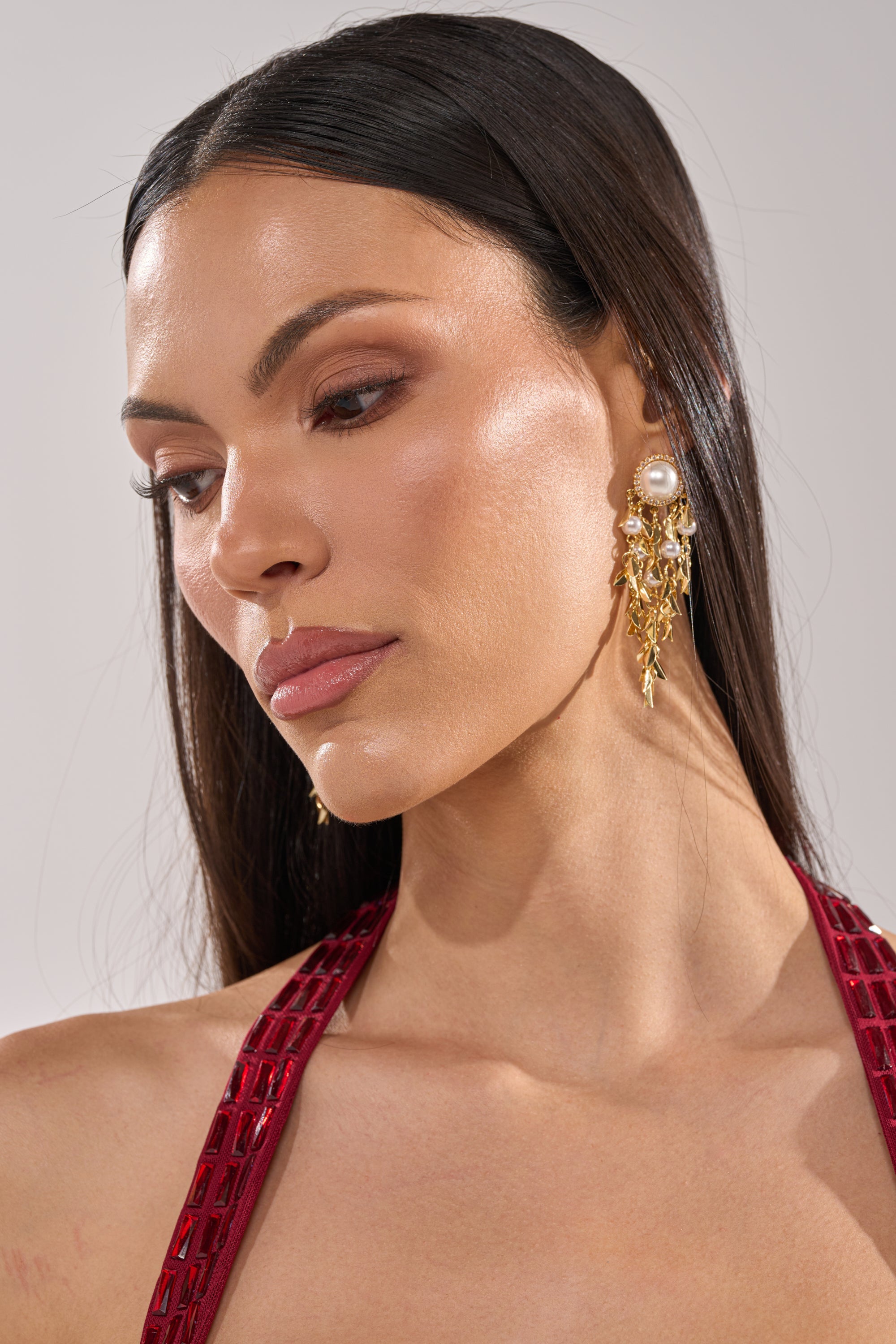 A woman with long dark hair wears the PARADISE EARRING—ornate gold and pearl statement jewelry—and a red textured dress. With natural makeup, she gazes downward serenely against a neutral background. 2