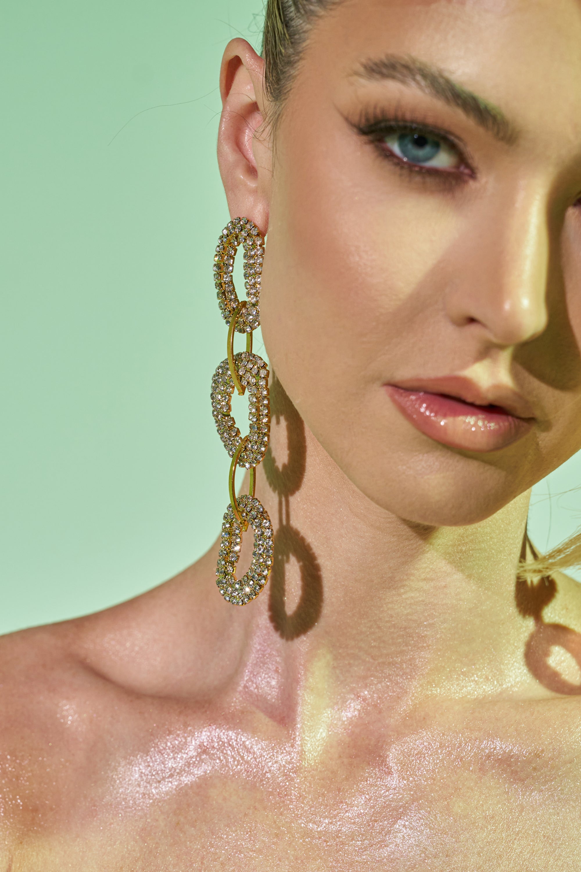 A close-up of a woman with glowing skin, blue eyes, and glossy lips wears the WILD ONE EARRING—crystal link earrings with three sparkling, connected oval loops—set against a green background. 0