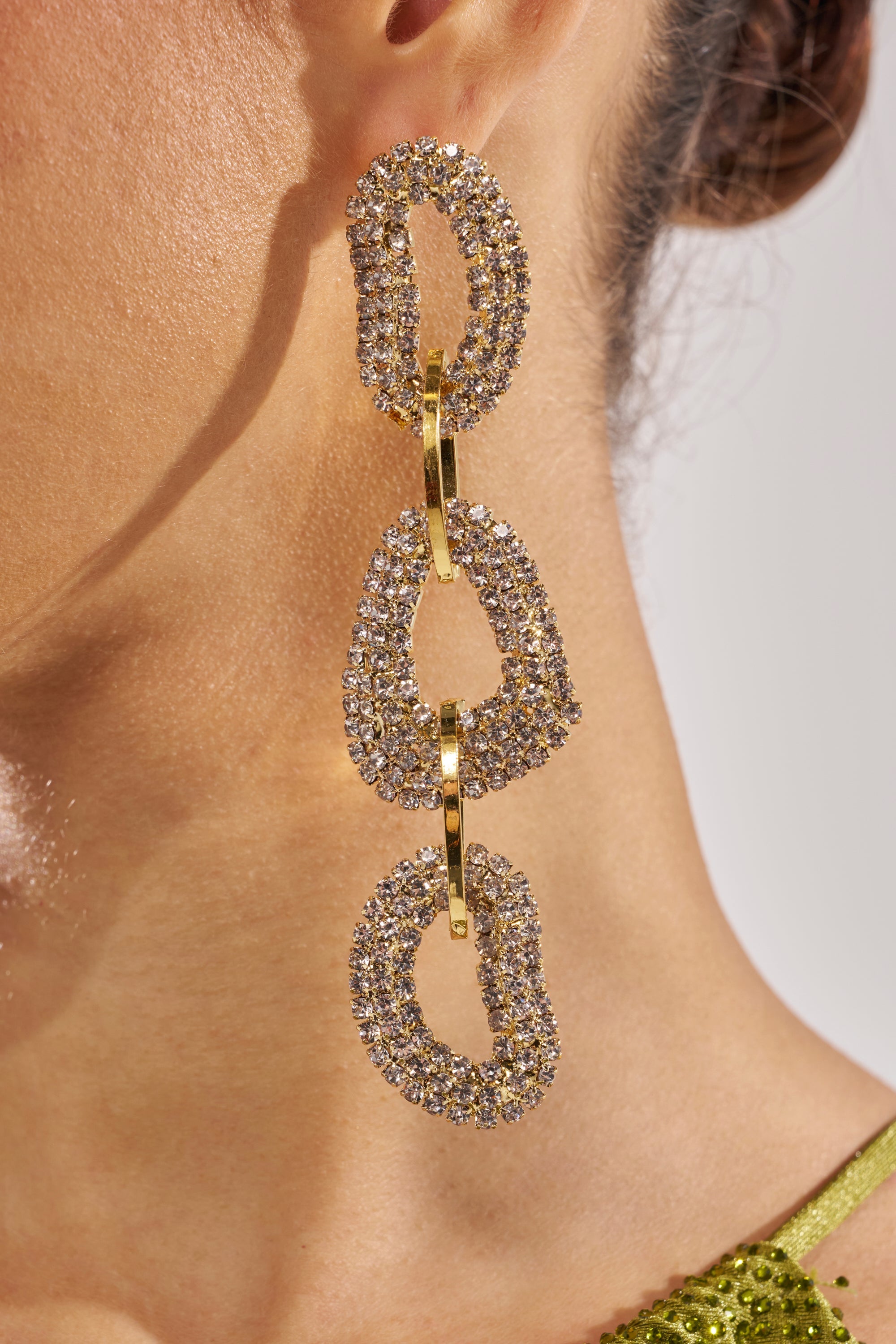 Close-up of a person wearing the WILD ONE EARRING, a large gold-tone, crystal-studded statement piece sparkling under the light. Their ear, jawline, and green top are visible. 3