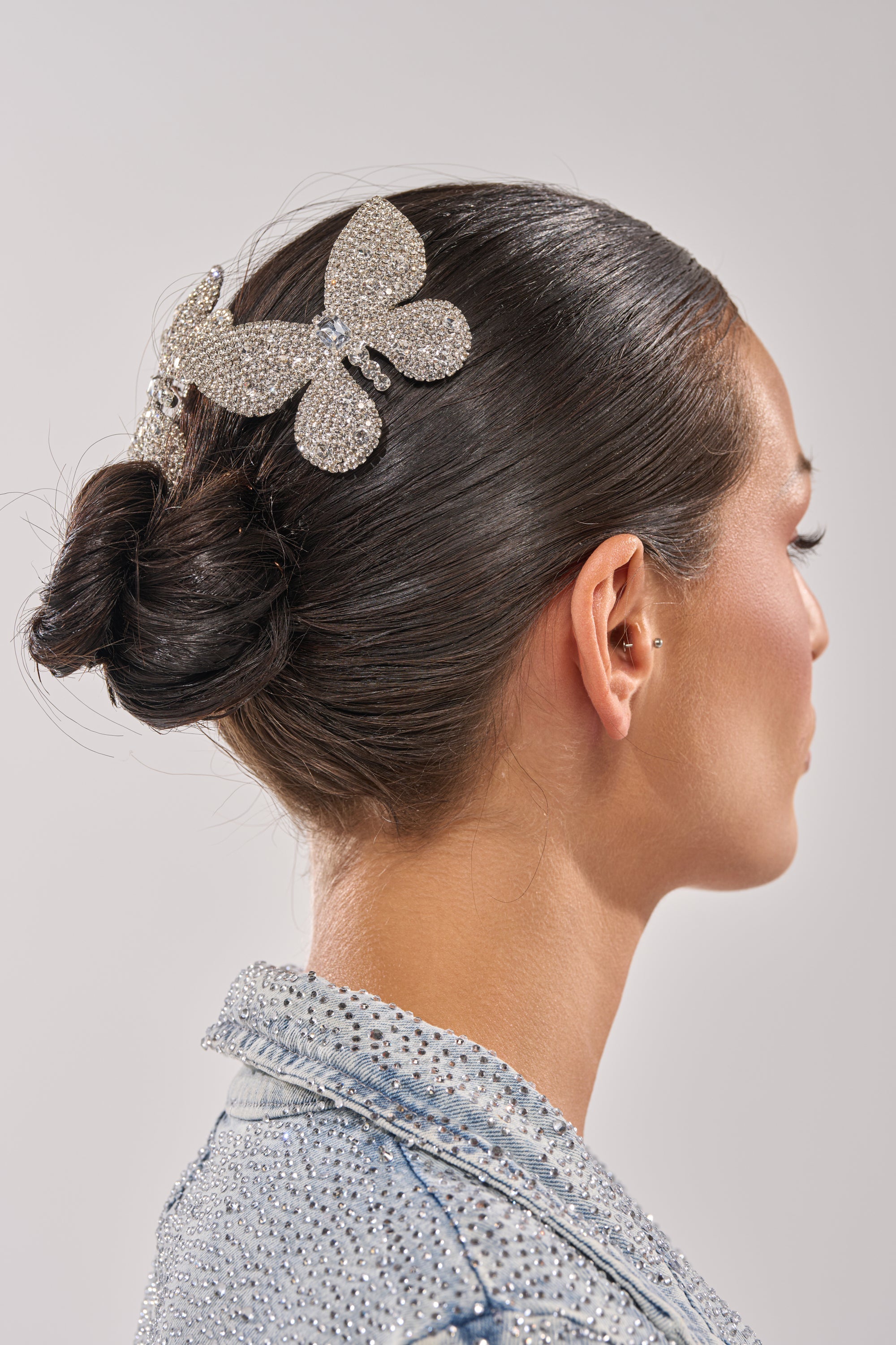 A woman with dark hair in a sleek bun wears a sparkling FLY AWAY HAIR CLIP SET and a silver, rhinestone-studded jacket. She is shown in profile against a plain background. 0