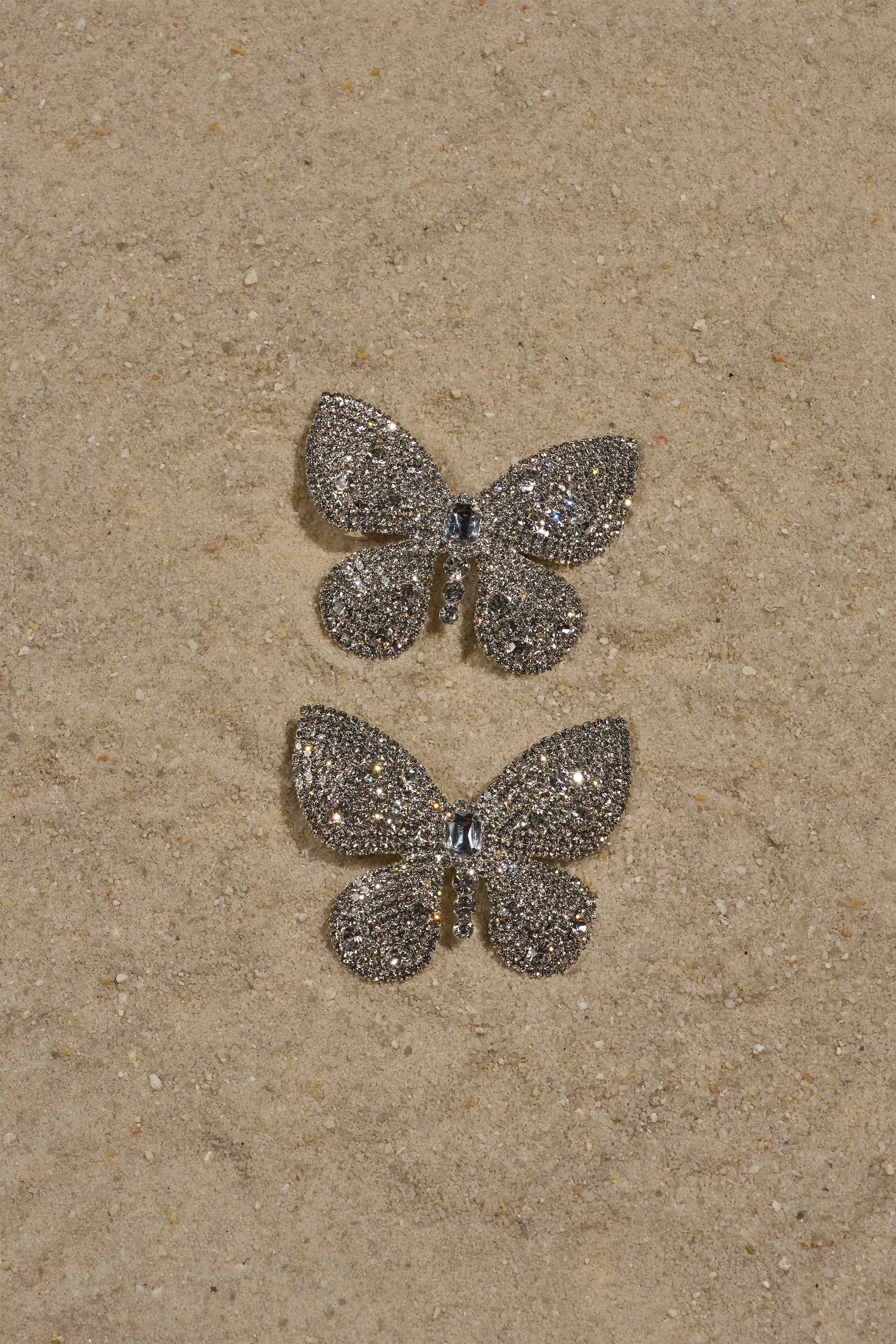 Two sparkling, silver butterfly-shaped clips from the FLY AWAY HAIR CLIP SET rest on sandy grains, catching the light and shining like crystal butterflies against the beige surface. 2