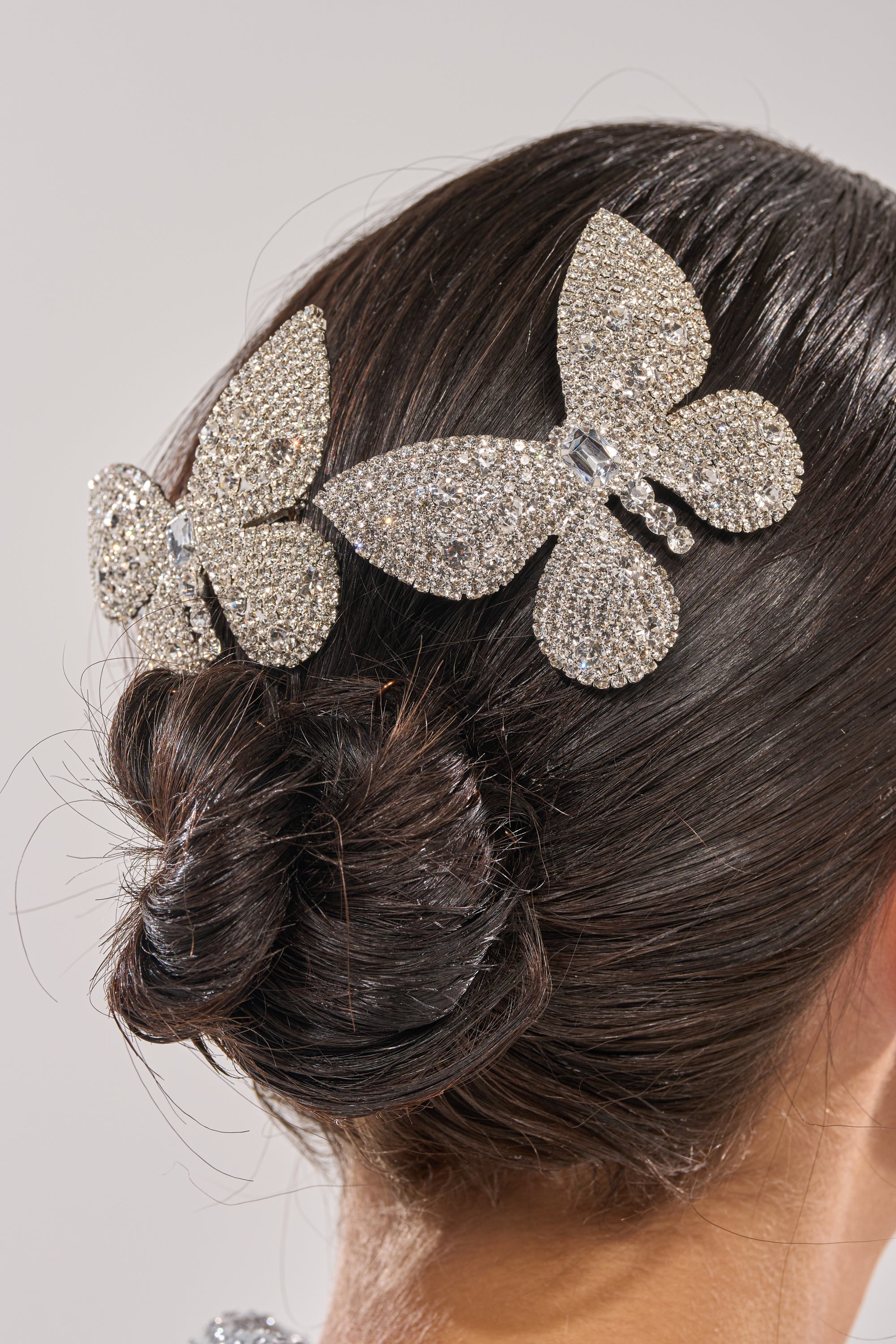 A close-up of a woman's dark hair in a low bun, embellished with two sparkling butterfly clips from the FLY AWAY HAIR CLIP SET. 1