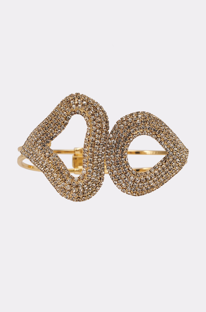 The ISNT THAT SWEET BRACELET is a gold rhinestone cuff with two abstract, overlapping loops adorned with sparkling clear crystals, showcased against a plain white background. 1