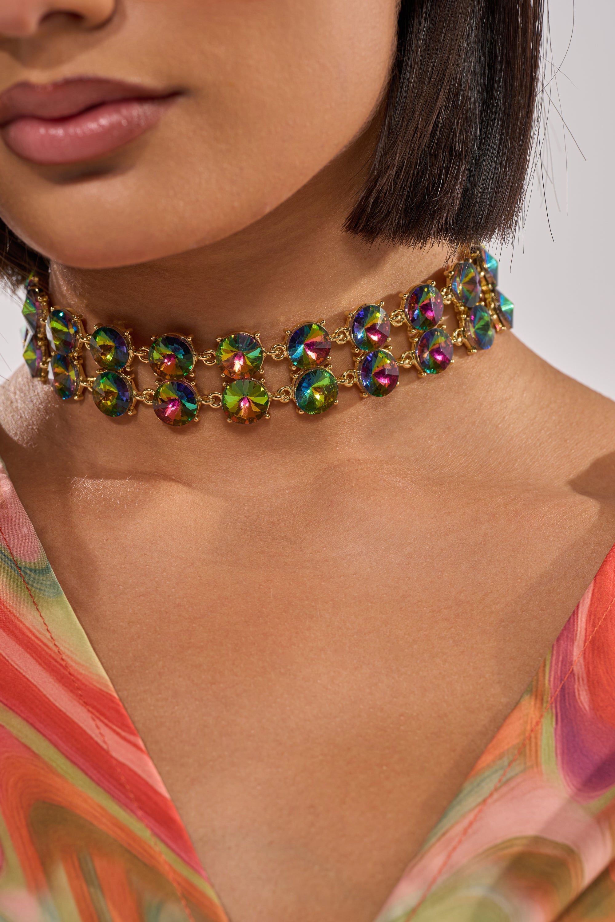 A woman with a bob haircut wears the U HAVENT SEEN THIS BEFORE NECKLACE, featuring colorful iridescent rhinestones in a gold setting and an adjustable extender chain. She sports light pink lipstick and a multicolored patterned top. 1