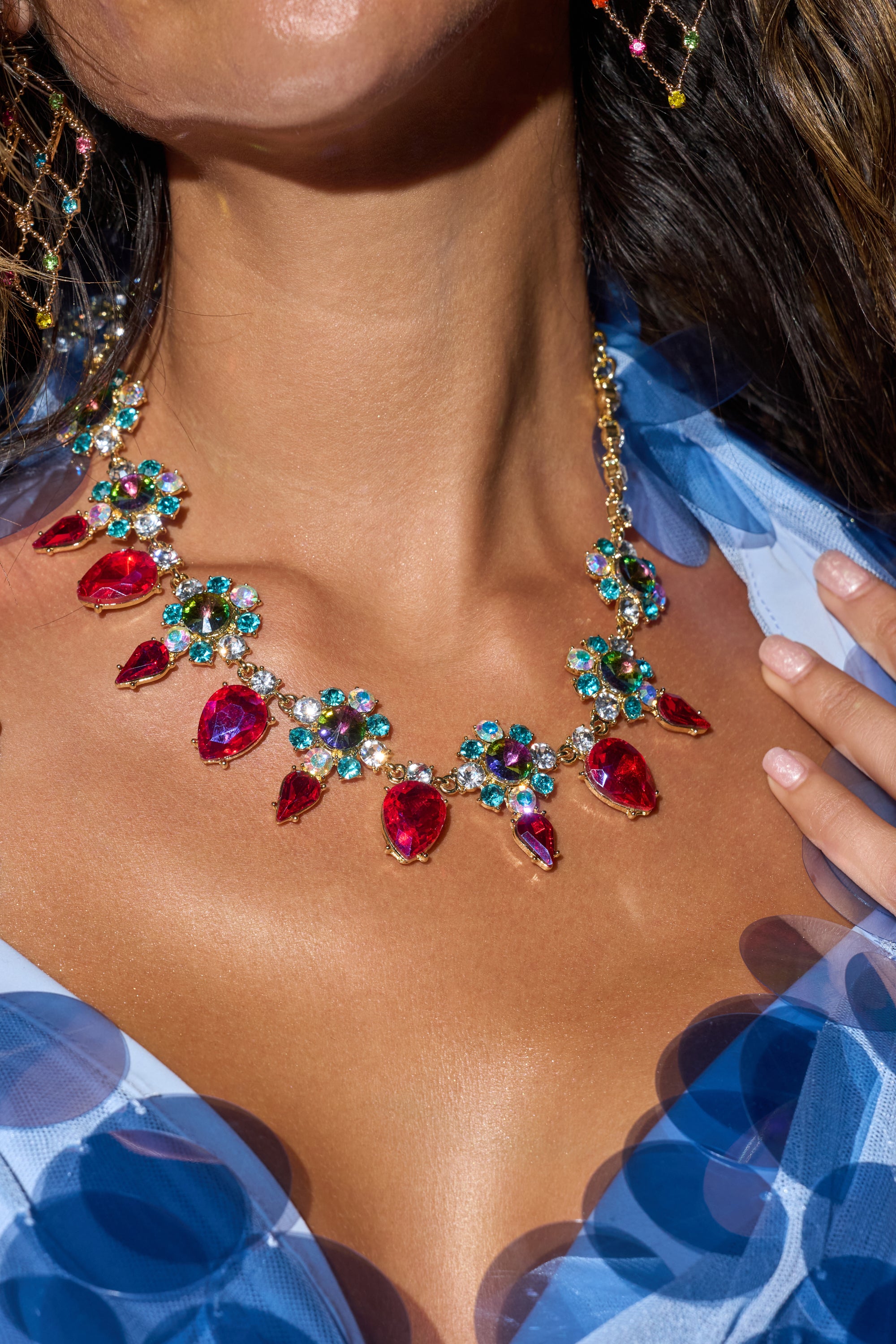 A close-up of a woman in a sheer blue dress, gently touching her collarbone while wearing the FESTIVAL NECKLACE—an eye-catching statement piece featuring large red gemstones and colorful crystal clusters.