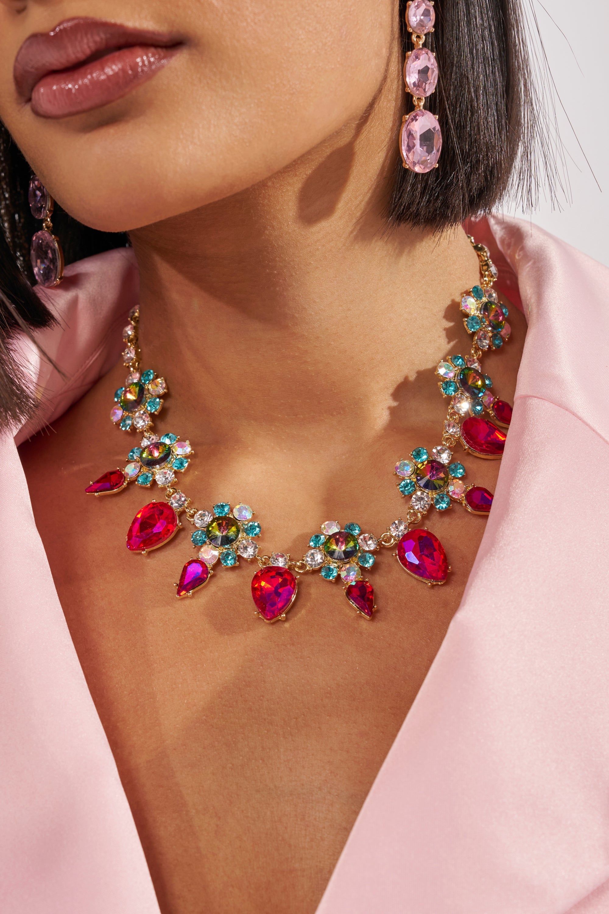 A woman in a light pink satin outfit showcases the FESTIVAL NECKLACE, featuring large red gemstones, multicolored crystals, and rhinestone accents, paired with matching pink gemstone drop earrings. Only the lower half of her face is visible. 1