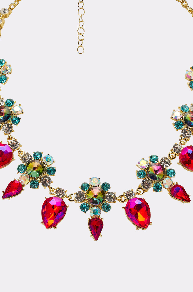 The FESTIVAL NECKLACE features bold red teardrop gems and sparkling floral clusters of green, blue, and clear crystals set in gold-tone metal, showcased on a plain background. 3