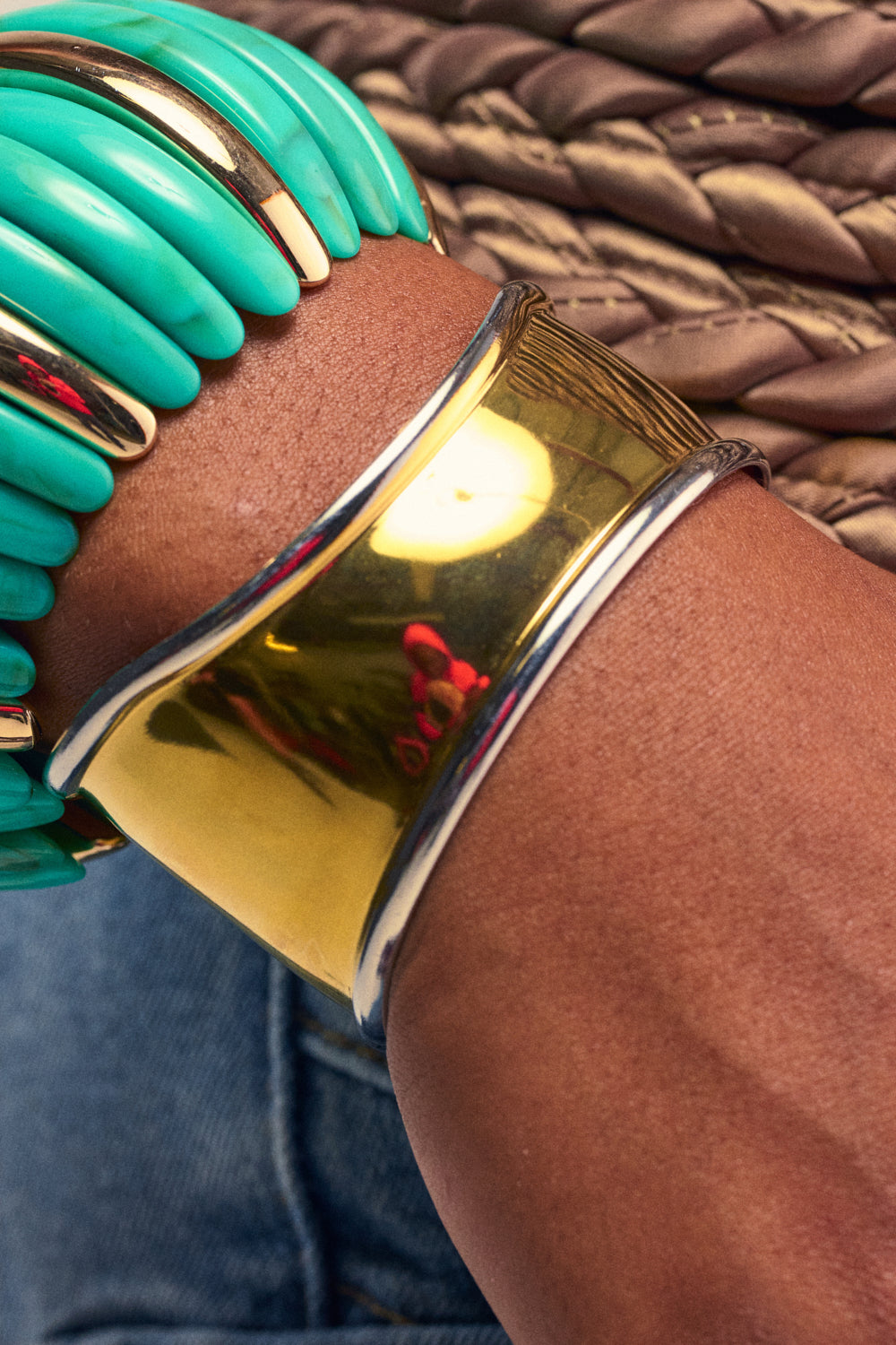 A close-up of a wrist wearing the CUFFING SZN BRACELET, styled with a chunky turquoise beaded bracelet. Shiny gold-tone metal gleams beside vibrant beads, while woven fabric and denim create a dynamic background. This chic stack highlights the CUFFING SZN BRACELET’s artful mix of textures and colors, offering an effortlessly cool take on mixed metal jewelry.