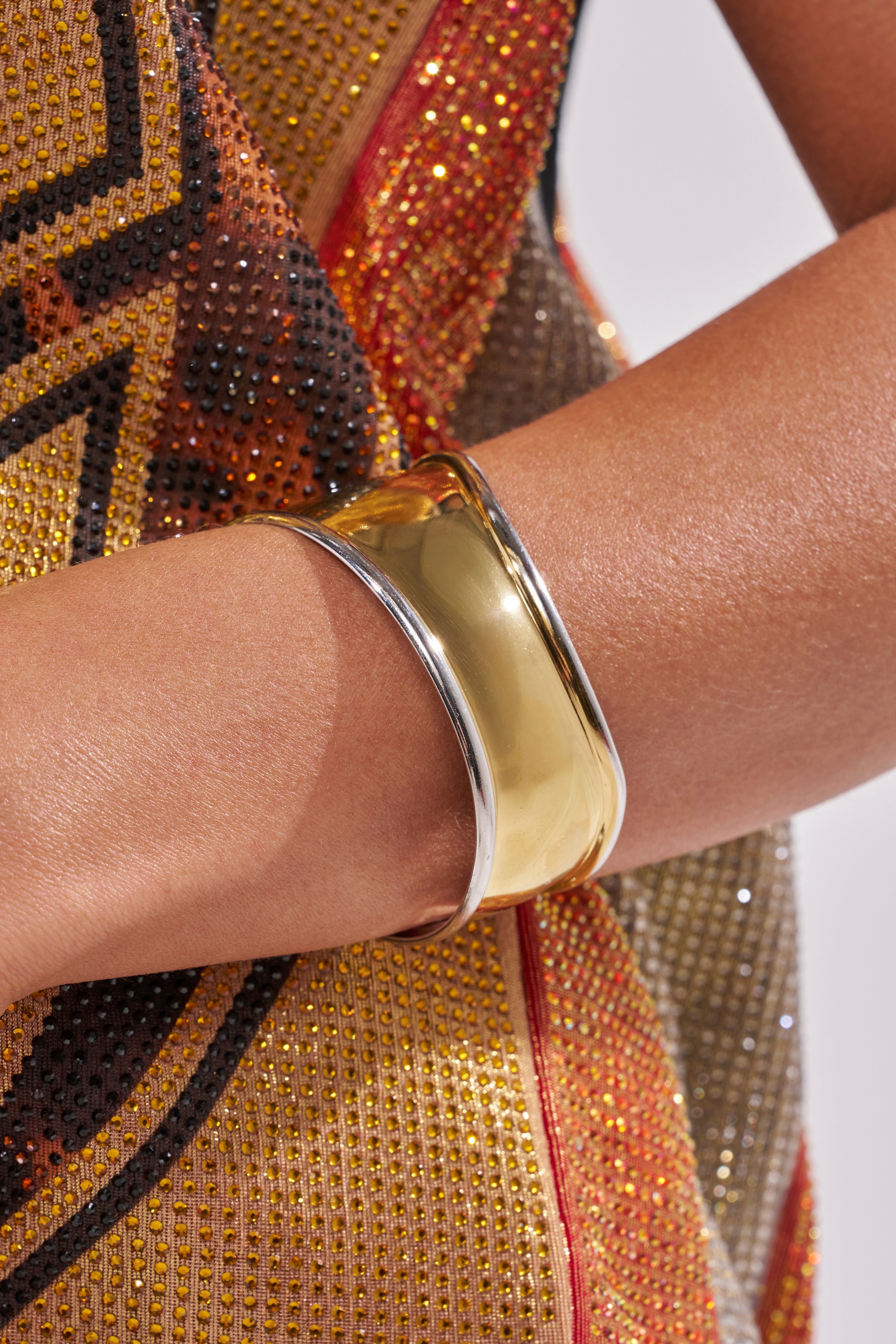 A close-up of a person's arm adorned with the CUFFING SZN BRACELET, a bold mixed metal cuff in shiny gold and silver, set against a sequined multicolored garment featuring detailed gold, red, and black patterns. 1