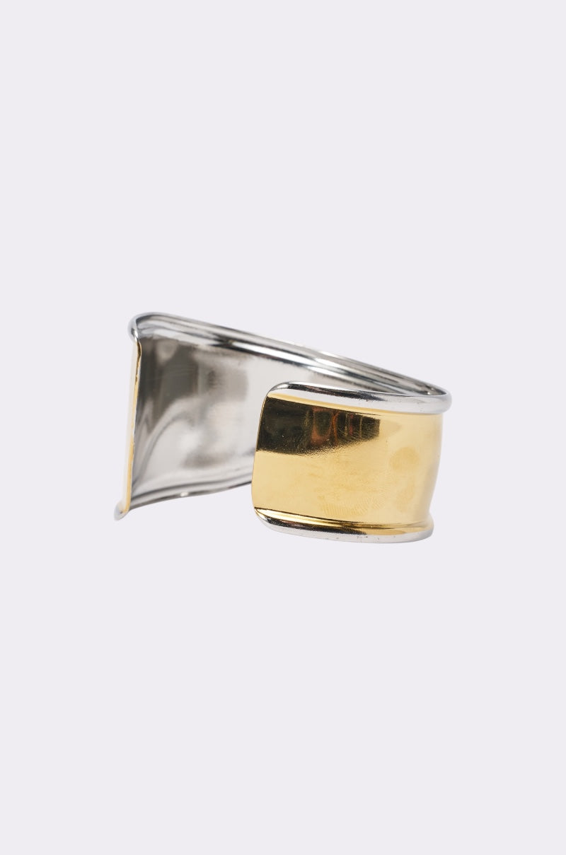 The CUFFING SZN BRACELET features a wide, open-ended sculptural cuff with a silver base and a bold gold-tone center, highlighted against a plain light gray background. This eye-catching piece embodies the appeal of mixed metal jewelry. 2