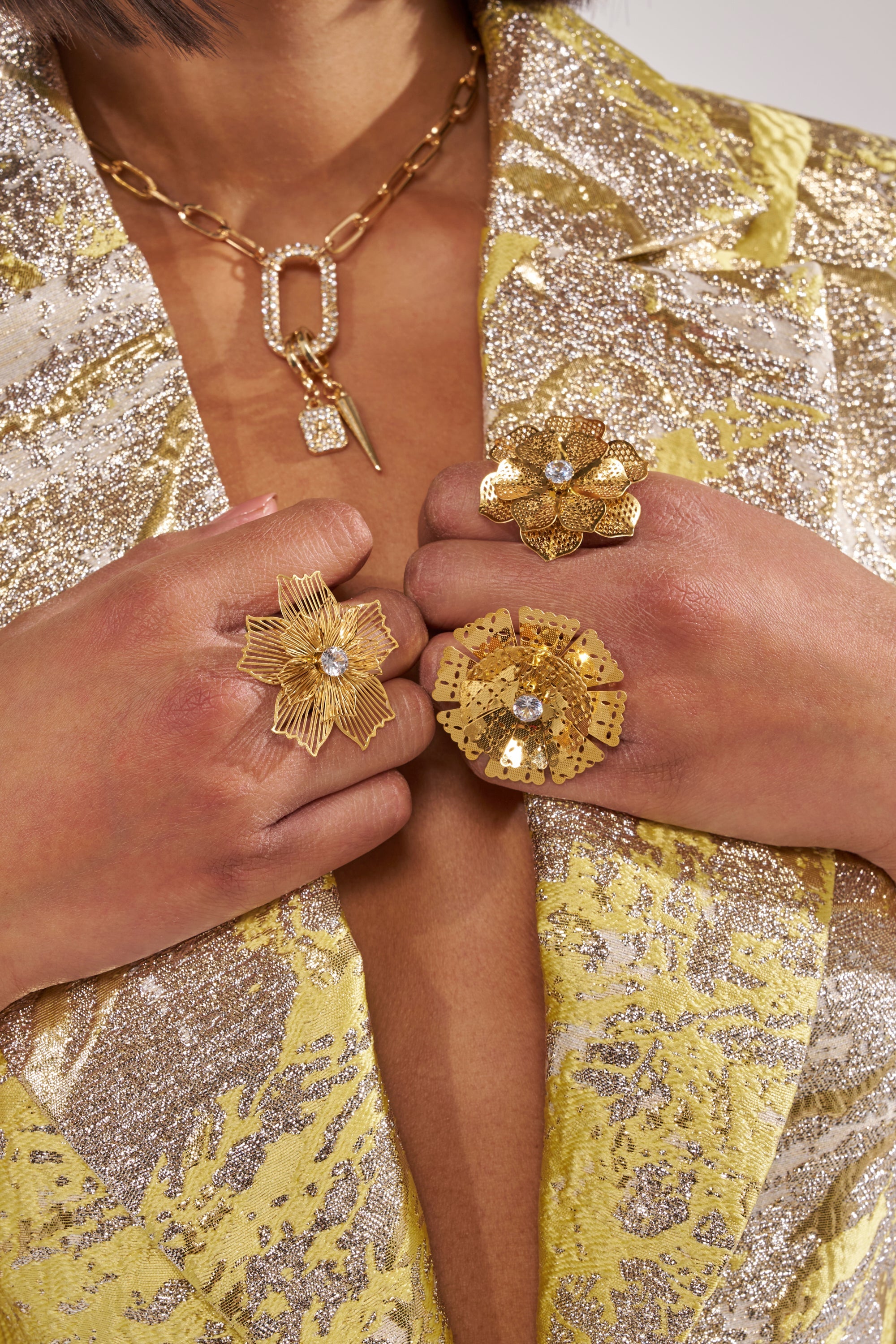 Wearing a gold and silver textured blazer, a person displays the LETS PARTY RING SET, featuring floral gold rings with adjustable bands, paired with a chunky gold chain necklace with geometric pendants. 1