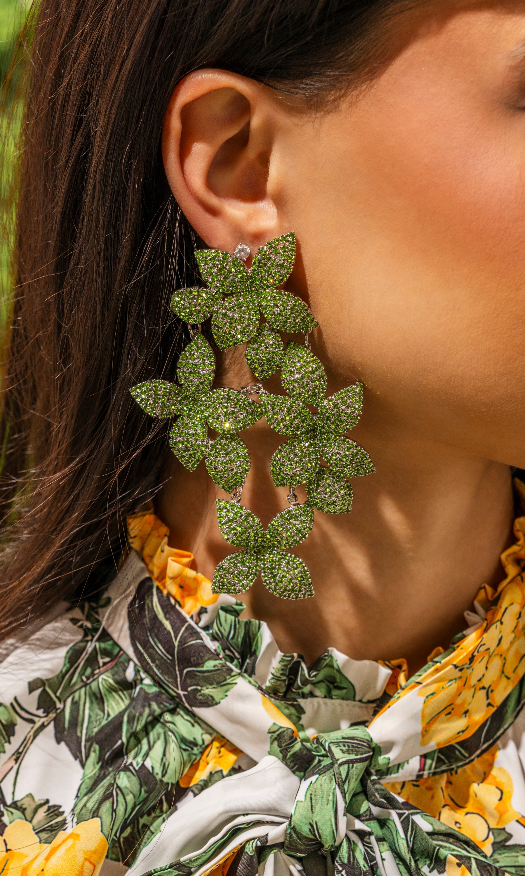A close-up of a woman wearing the bold IN MY GARDEN EARRING—large green drop earrings with sparkling rhinestones. She pairs them with a yellow and green floral blouse that highlights the statement earrings. 0