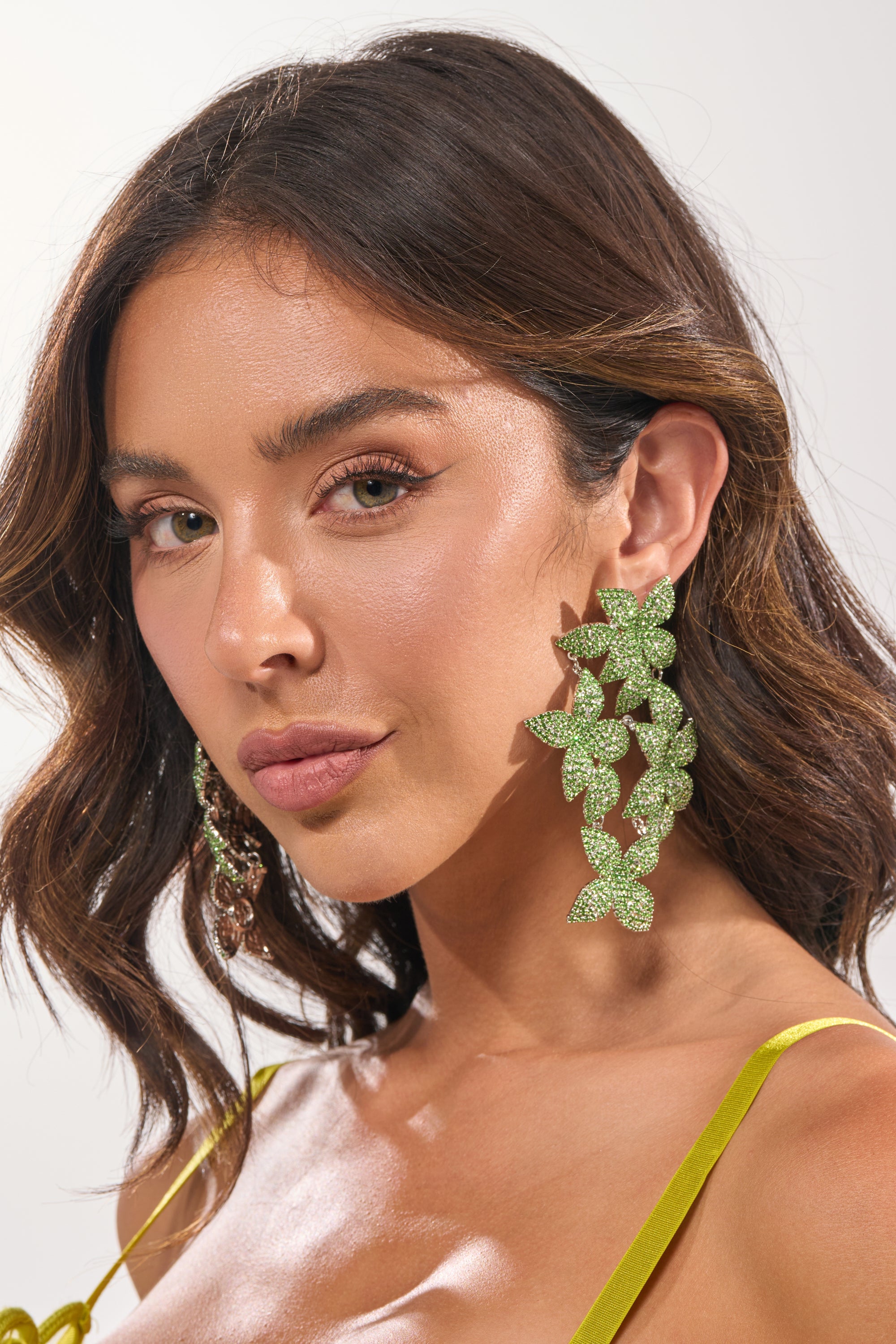 A woman with wavy brown hair wears the IN MY GARDEN EARRING—green, ivy leaf-shaped drop earrings—and a yellow spaghetti strap top, confidently facing the camera against a neutral background. 2