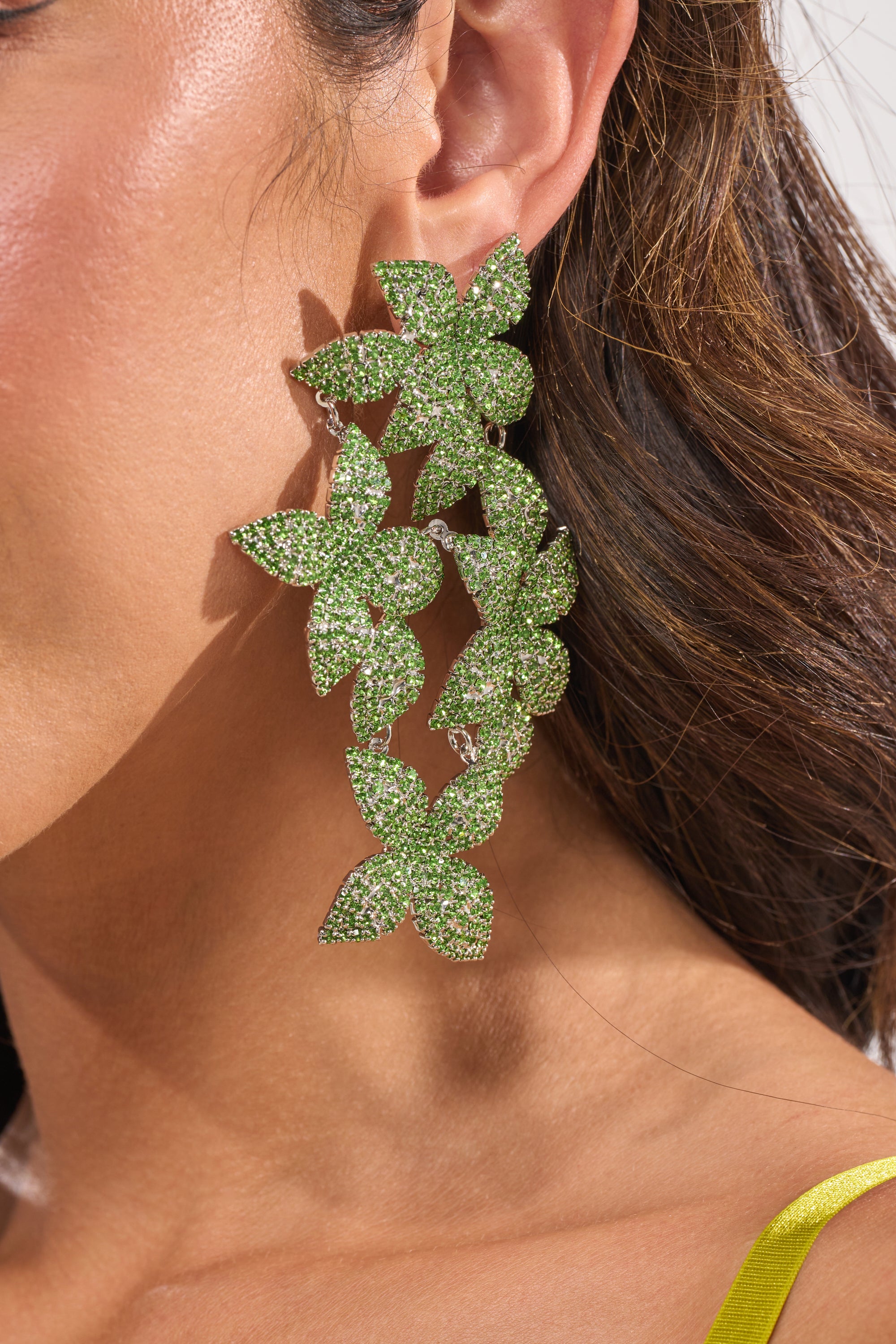Close-up of a woman's ear wearing the IN MY GARDEN EARRING, featuring bold green floral rhinestones. The tiered statement earrings sparkle and stand out against her brown hair and yellow clothing strap. 3