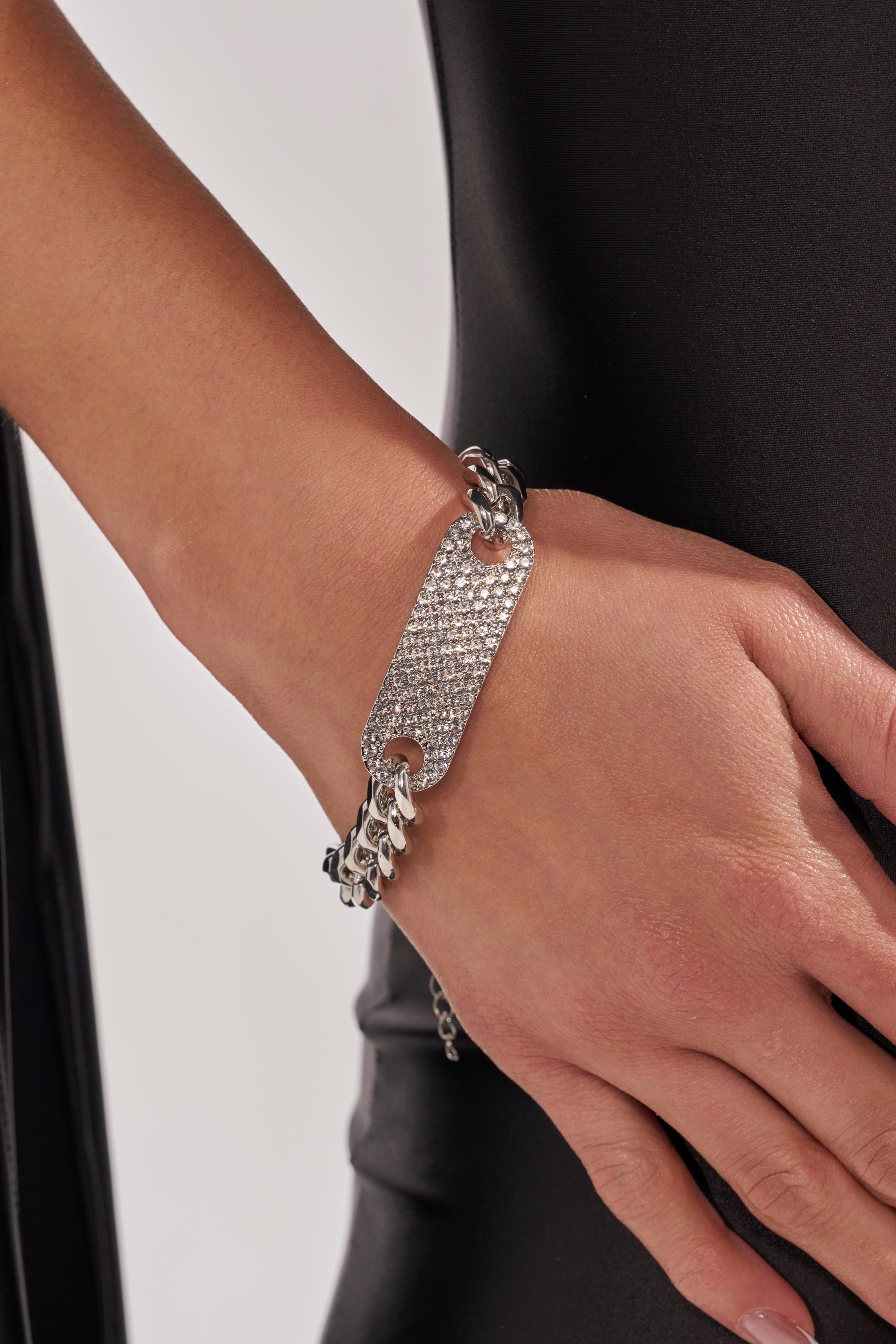 A close-up of a person wearing the LETS LINK BRACELET, a chunky ID bracelet with a rectangular, rhinestone-encrusted plate, resting on their hand against a black outfit. 1