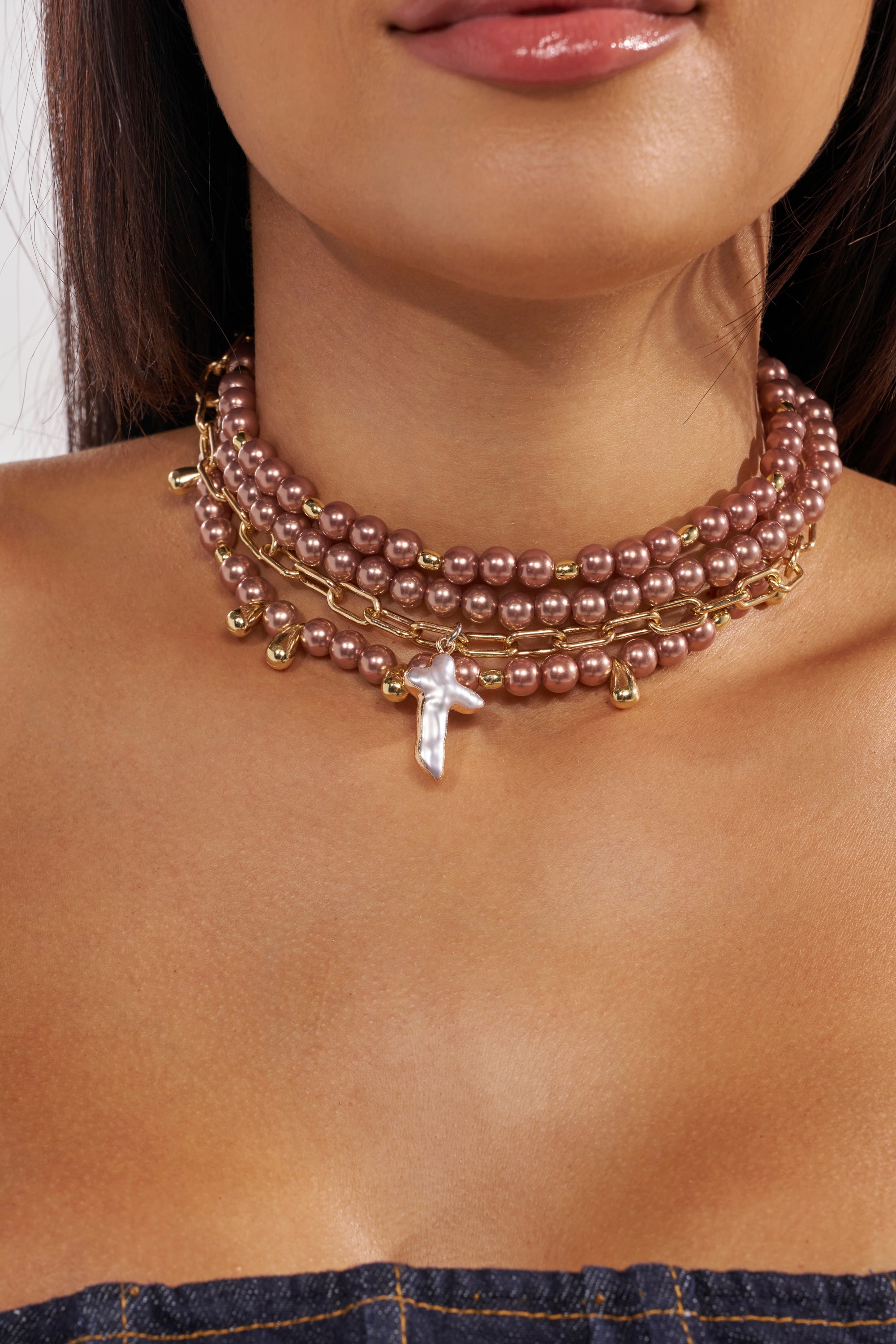 A woman wears the TAKE IT THERE NECKLACE, a multi-layered choker of pink and gold beads with a faux pearl cross pendant, her face and shoulders partly visible. 1