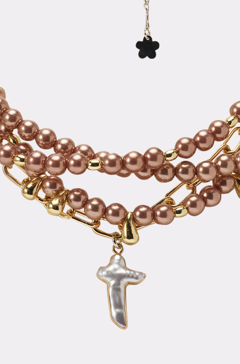 The TAKE IT THERE NECKLACE features three strands of shiny bronze faux pearls, a gold paperclip chain, a gold-edged pearlescent cross pendant at the center, and a delicate black flower charm above. 2