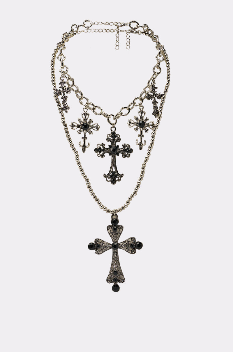 The IN THESE STREETS NECKLACE showcases five ornate black cross pendants of different sizes and intricate designs, layered on multiple silver chains, all presented against a plain white background. 3