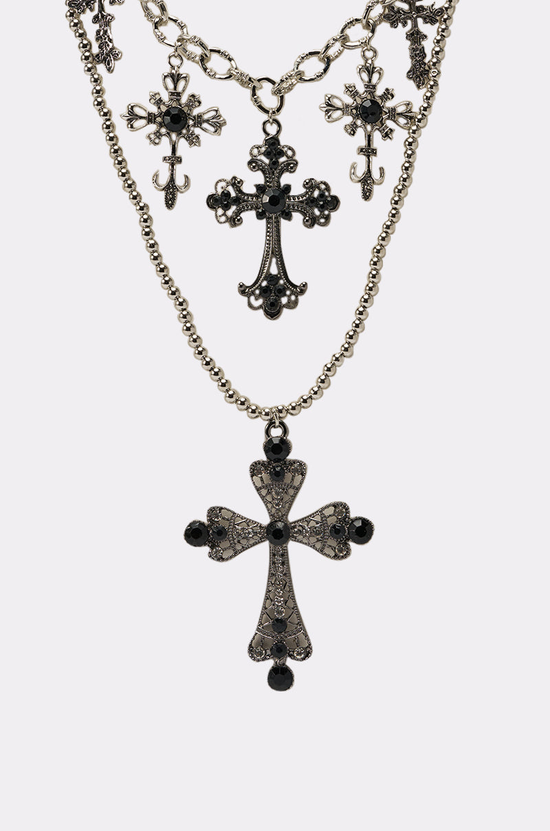 The IN THESE STREETS NECKLACE set showcases three layered silver cross pendants with black gem accents on a white background. The largest cross hangs from a beaded chain, while two smaller crosses feature unique textured chains above. 4