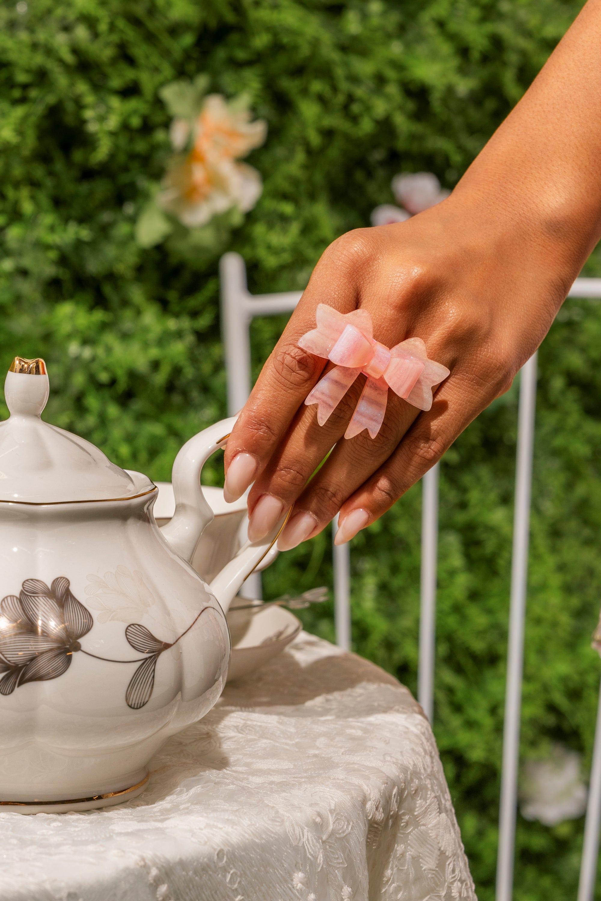A hand with long, manicured nails wears the KEKE PINK BOW RING and coquette jewelry while holding the lid of a white floral teapot on a lace-covered table, with flowers and green leaves in the background. 0