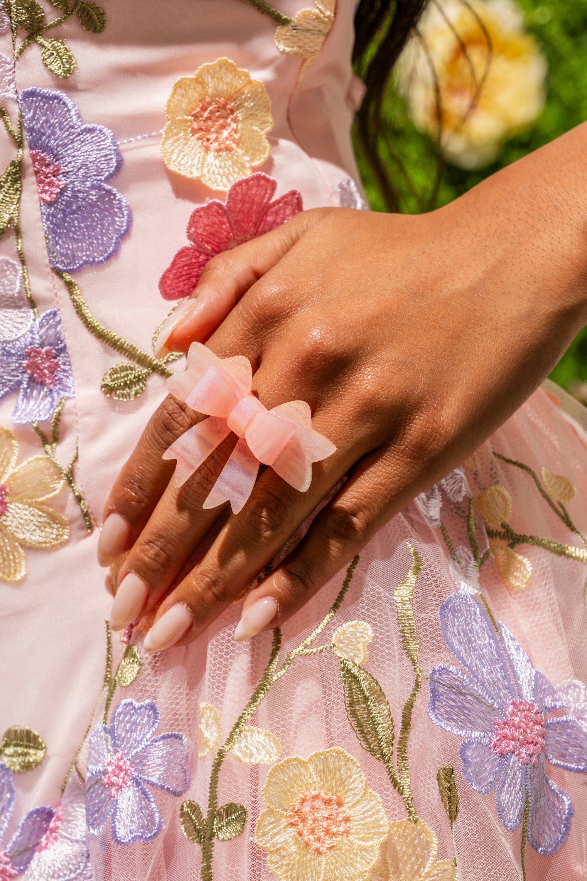 A hand with long, nude nails rests on a pink floral dress, wearing the KEKE PINK BOW RING—a large, pink marbled bow. Blurred green grass and yellow flowers are in the background. 1