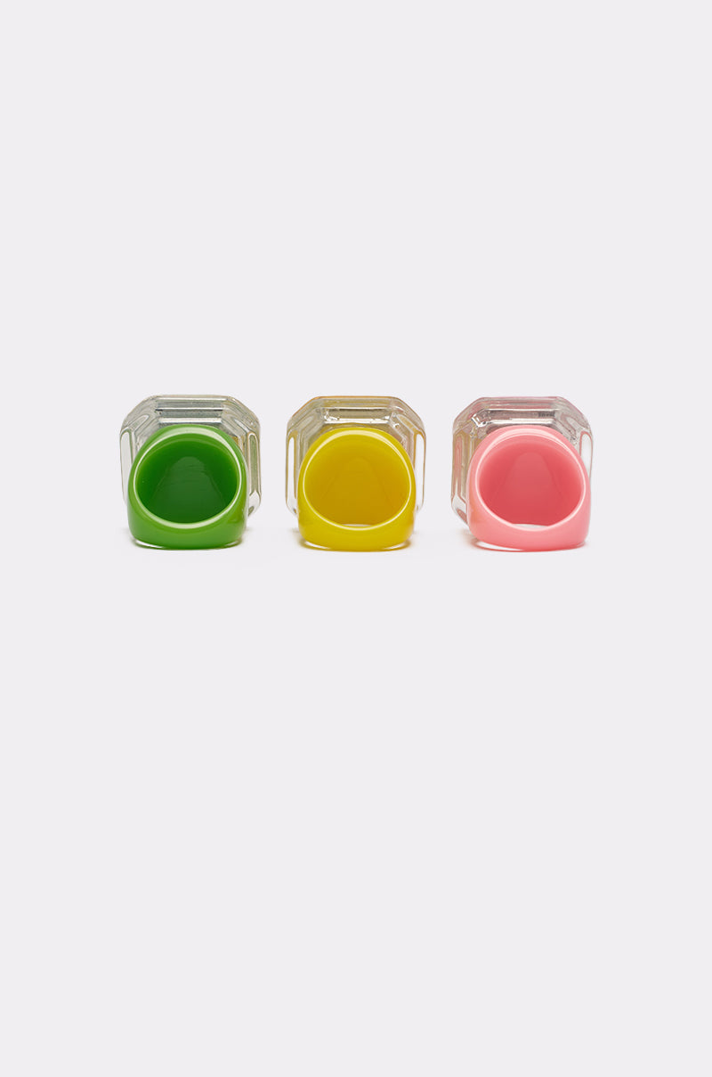 Three small glass jars viewed from above, each filled with green, yellow, and pink substances, are arranged in a row on a plain white background, reminiscent of the vibrant GLINDA VIBES RING SET's square-cut acrylic gem stones. 3