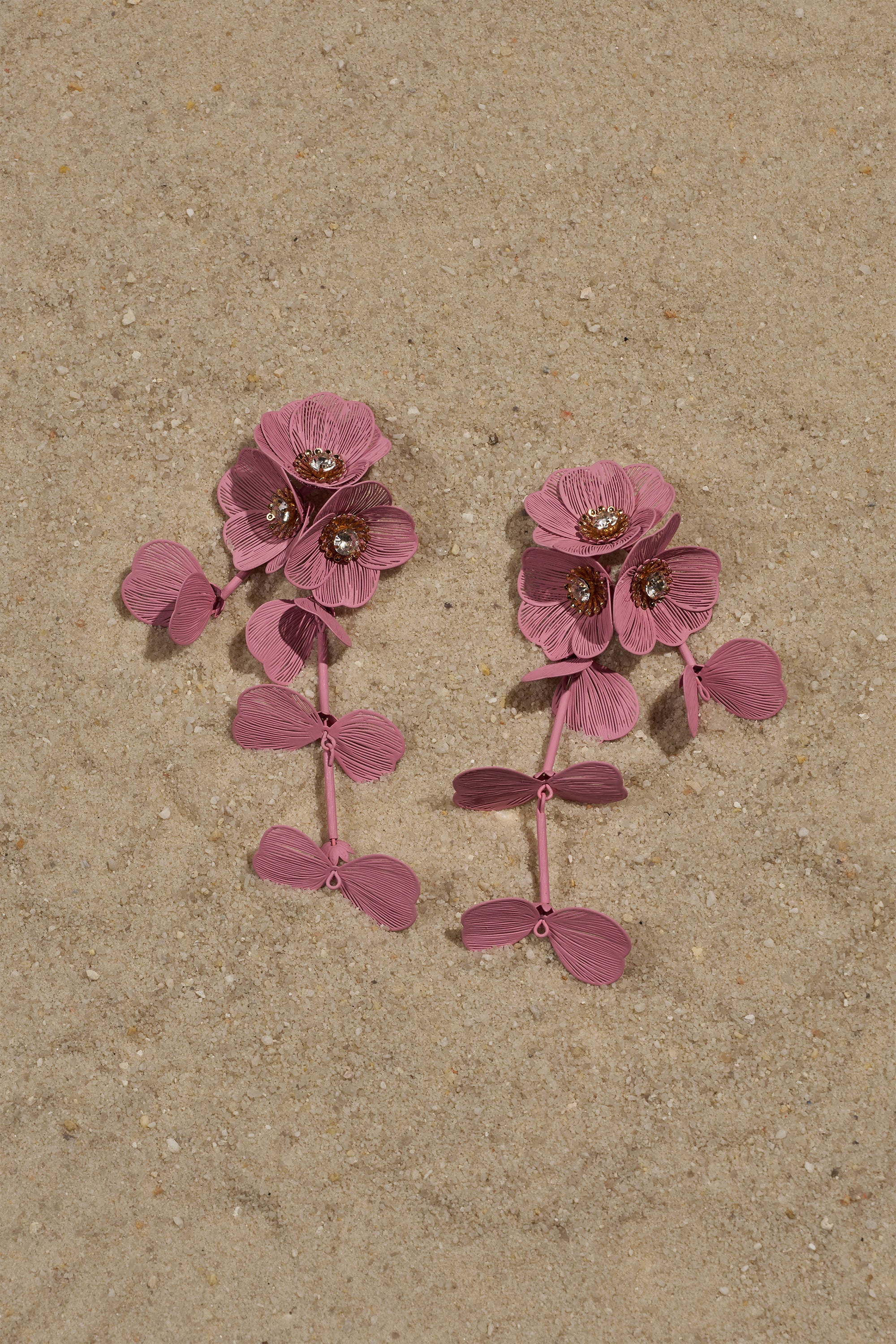 The DO TOO MUCH EARRING features pink, flower-shaped earrings with textured petals and gem centers, arranged on sandy ground. 1