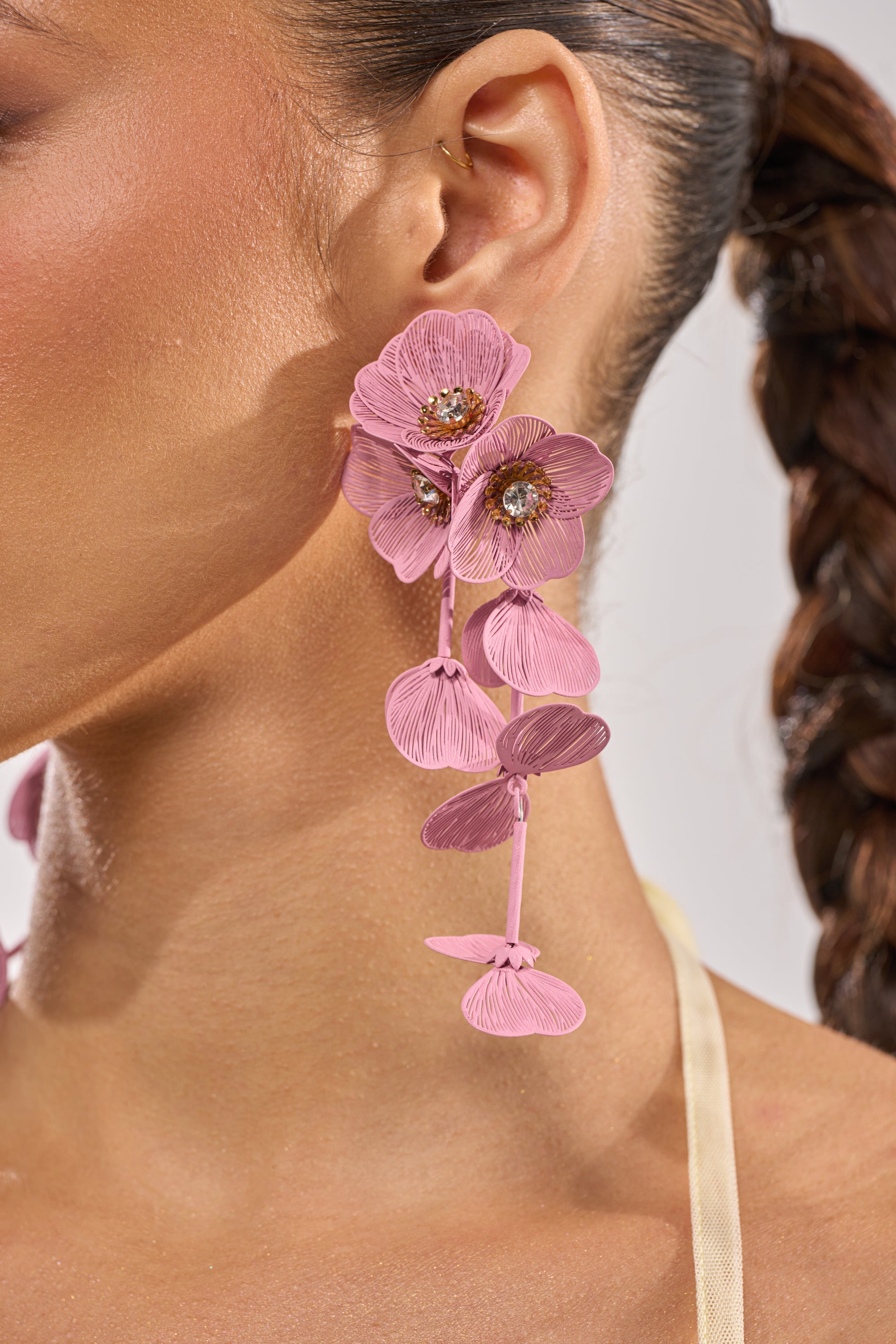 Close-up of a person wearing the DO TOO MUCH EARRING—large, intricate pink flower earrings with detailed petals and sparkling centers. Their braided hair highlights the crystal earrings against their warm skin tone. 4