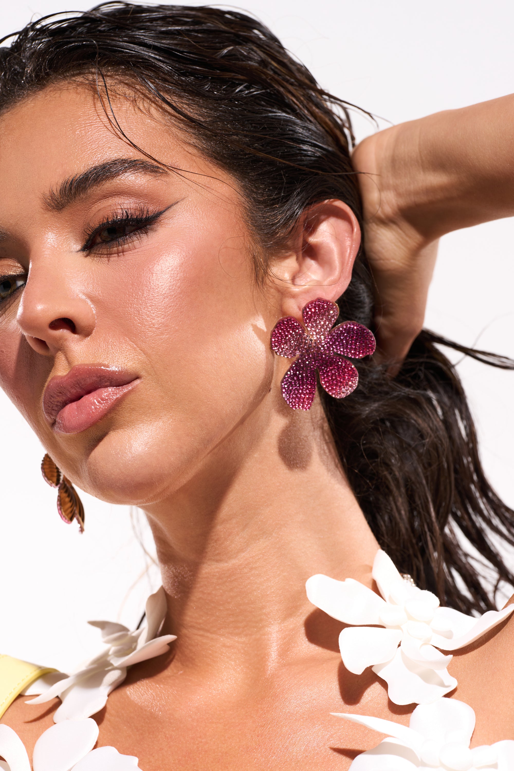 A woman with wet, slicked-back hair poses with her hand behind her head, wearing the WORTHY EARRING—large crystal finish earrings shaped like purple flowers—and a white floral necklace against a bright background. 0