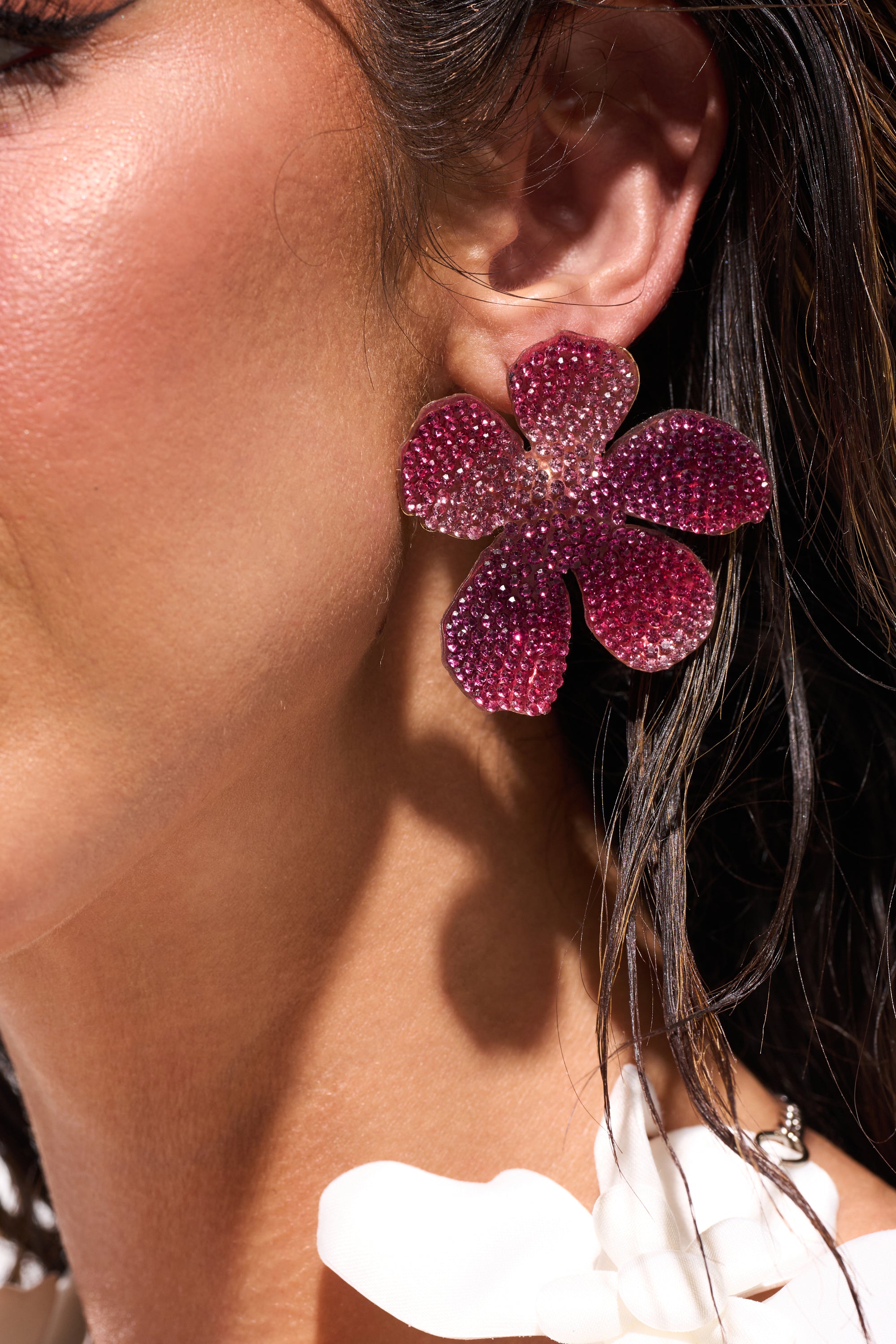 Close-up of a person wearing the WORTHY EARRING—a large, sparkling, purple floral stud. Their tanned skin and wet dark hair are visible, along with part of a white ruffled garment near the neck. 1