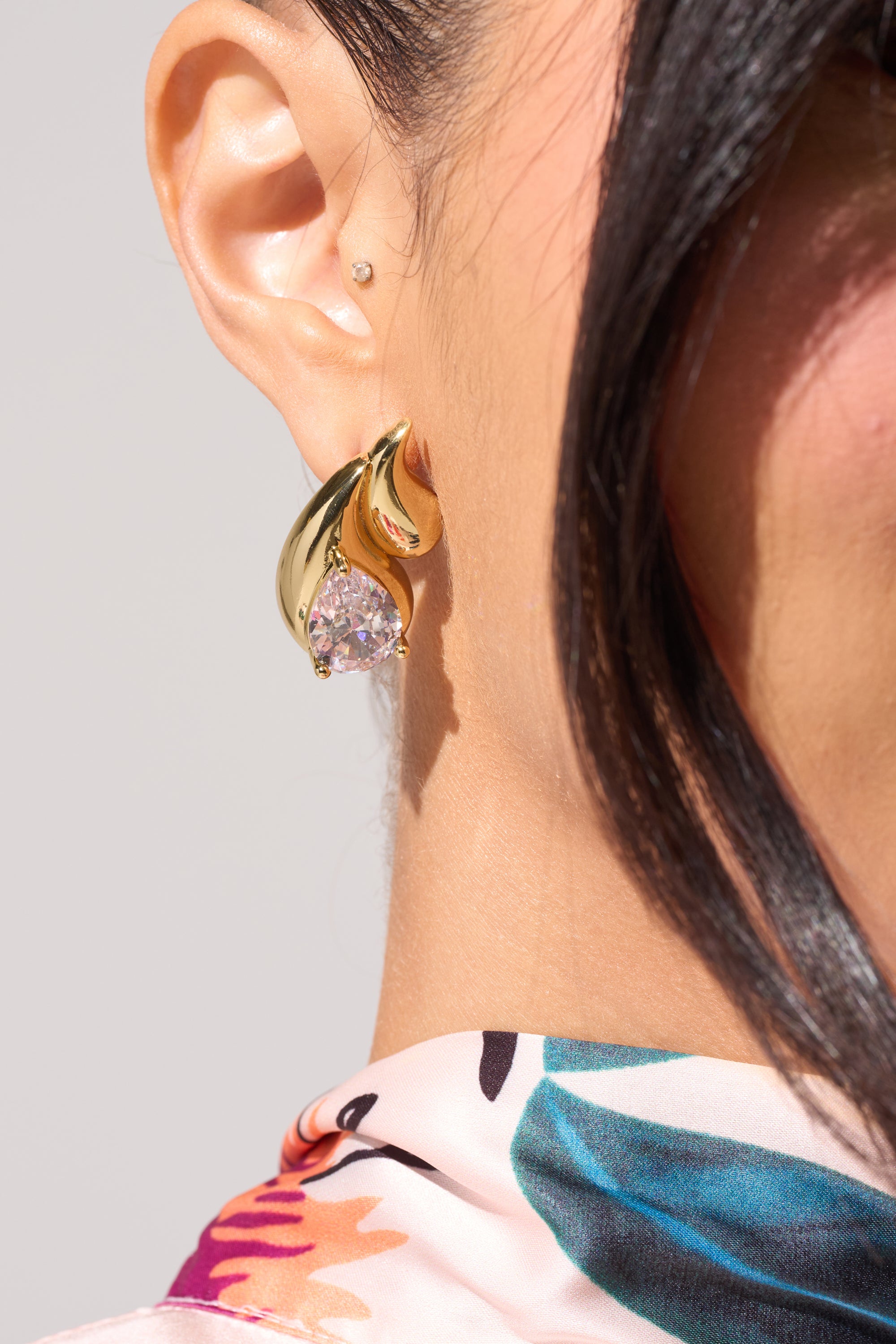 Close-up of a woman's ear wearing the OLD MONEY EARRING—a gold teardrop design—paired with a crystal stud and colorful patterned scarf, creating an elegant look perfect for wedding earrings inspiration. 0