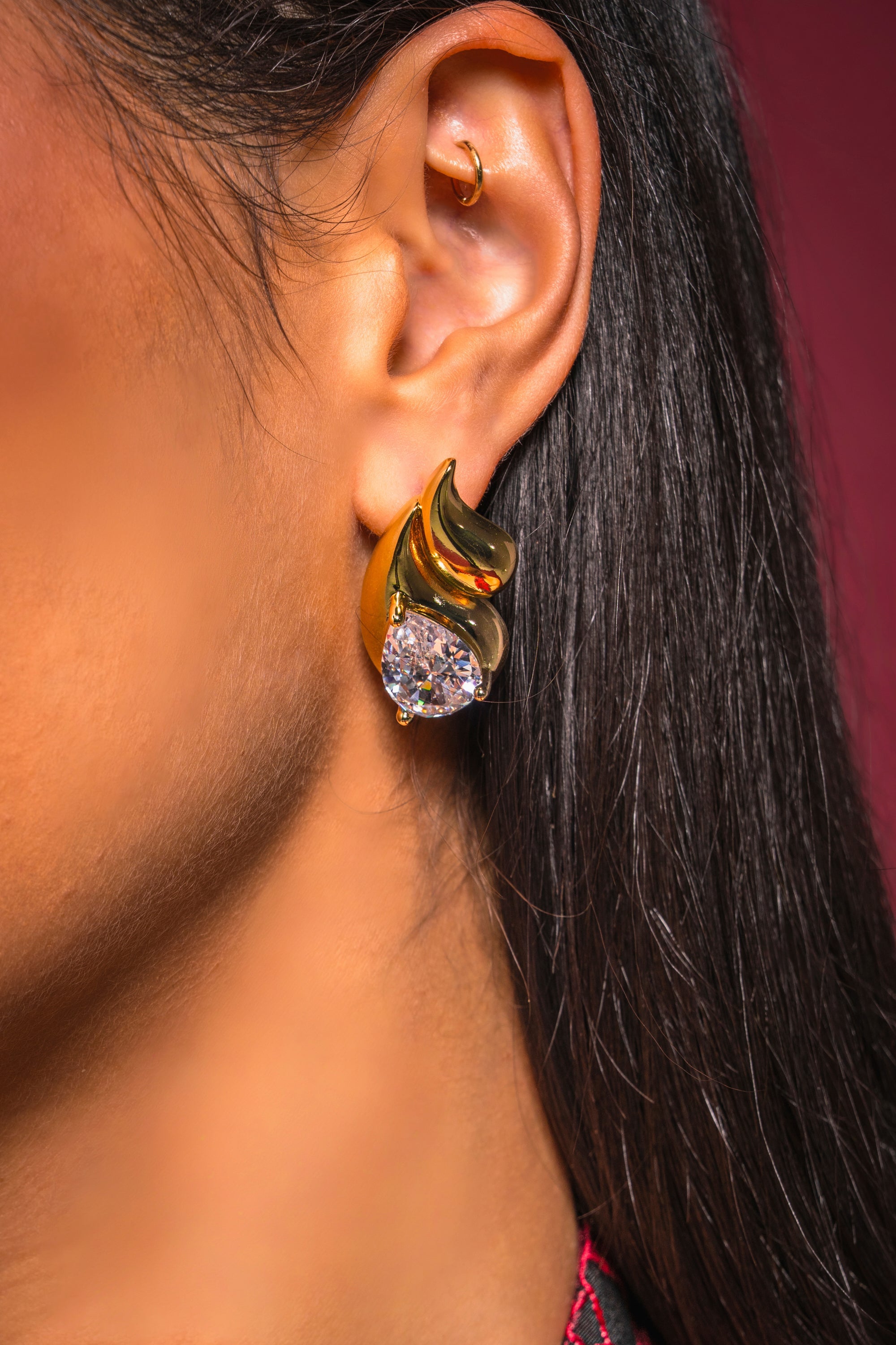 A close-up of a person’s ear wearing the OLD MONEY EARRING—a gold teardrop with a large, clear gemstone and a small hoop in the upper ear—stands out against a rich burgundy background, highlighting their elegant formal style. 2