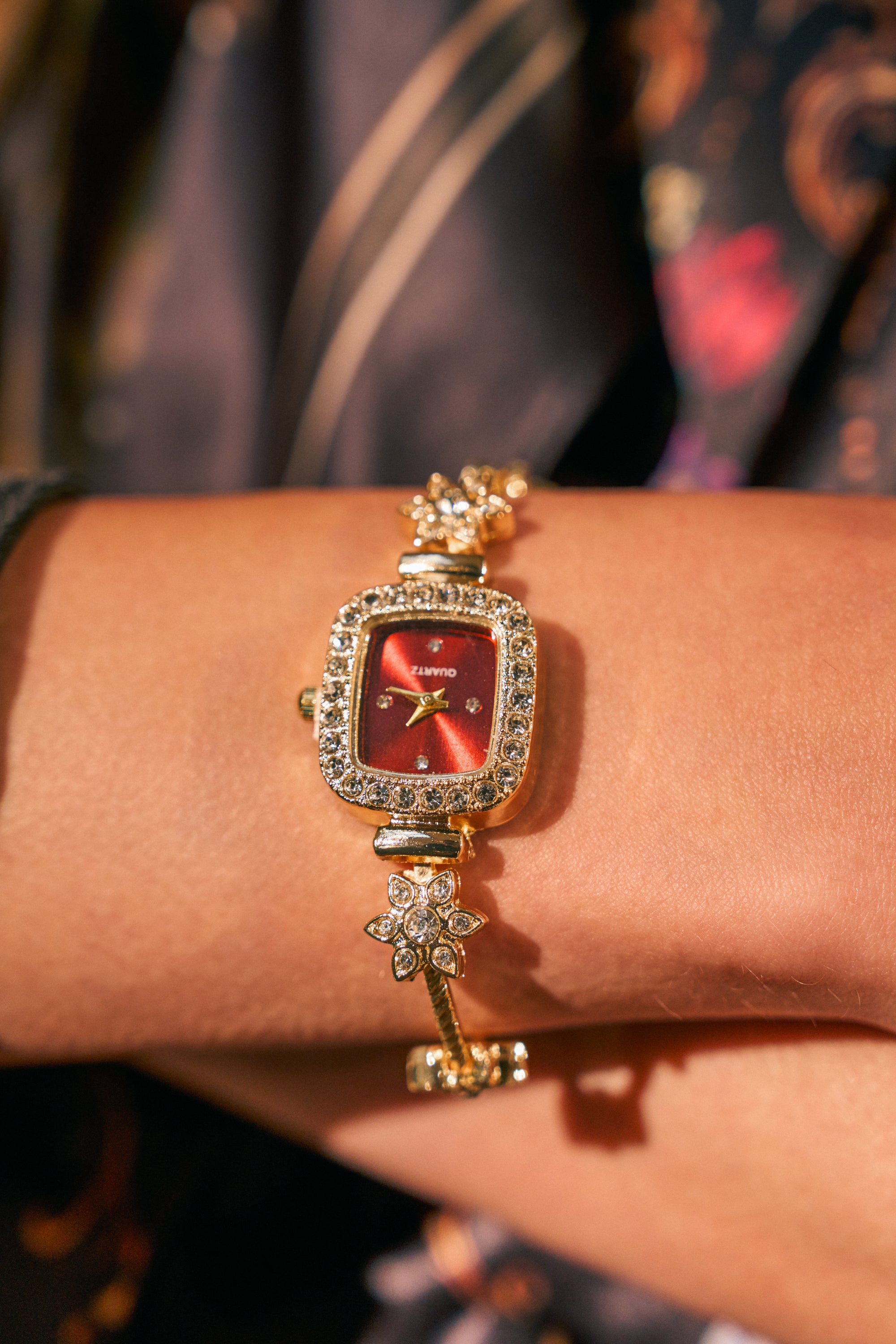 The ELEGANCE WATCH, featuring a gold bracelet band with floral details and ornate, vintage-inspired design, is worn on a person’s wrist. Its red square face is surrounded by small rhinestone accents, giving it a decorative and sophisticated appearance.