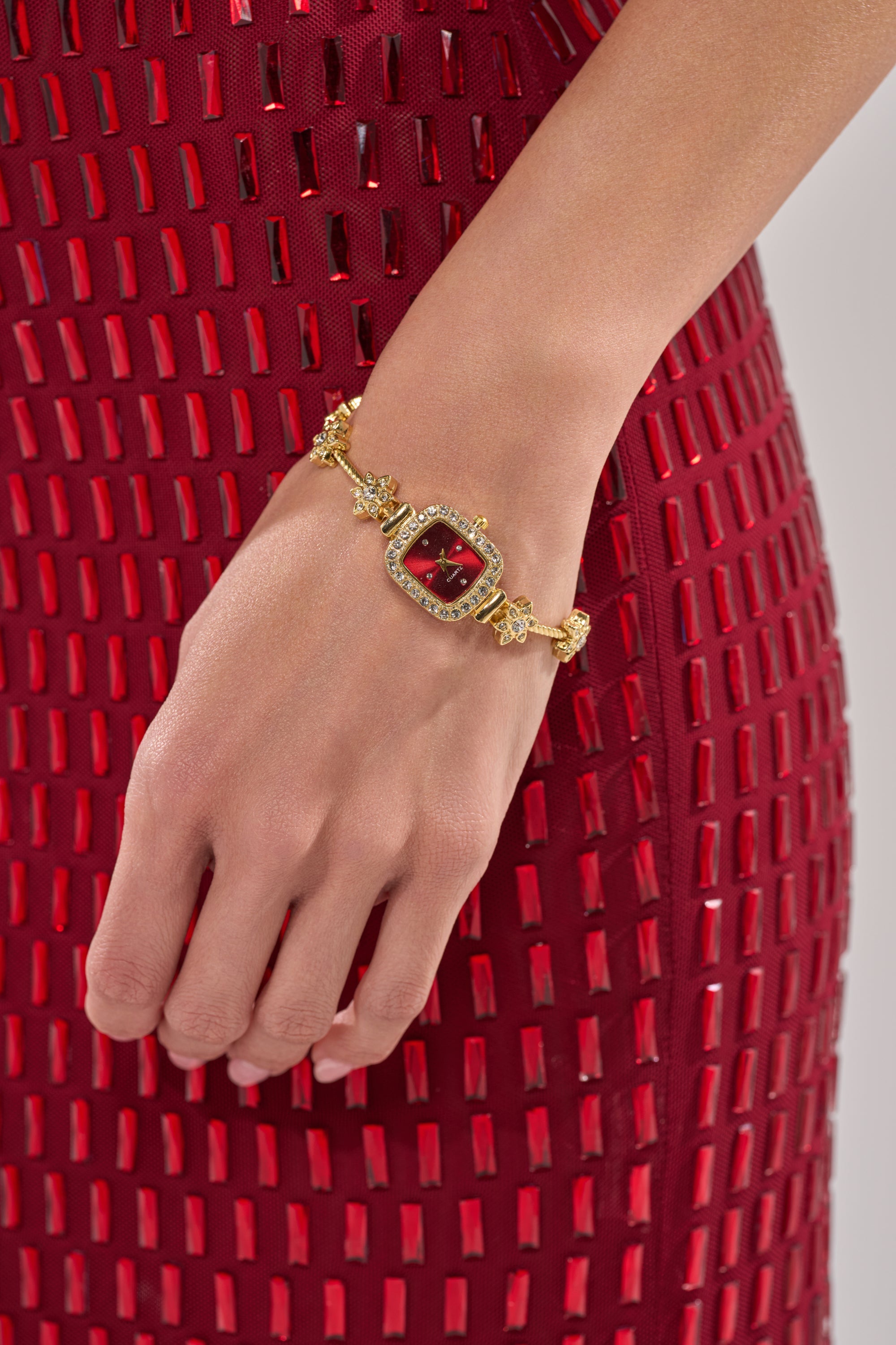 A close-up of a woman's hand wearing the ELEGANCE WATCH, a vintage-inspired gold bracelet watch with a red gemstone, rhinestone accents, resting against a red dress with geometric rectangular cutouts. 1