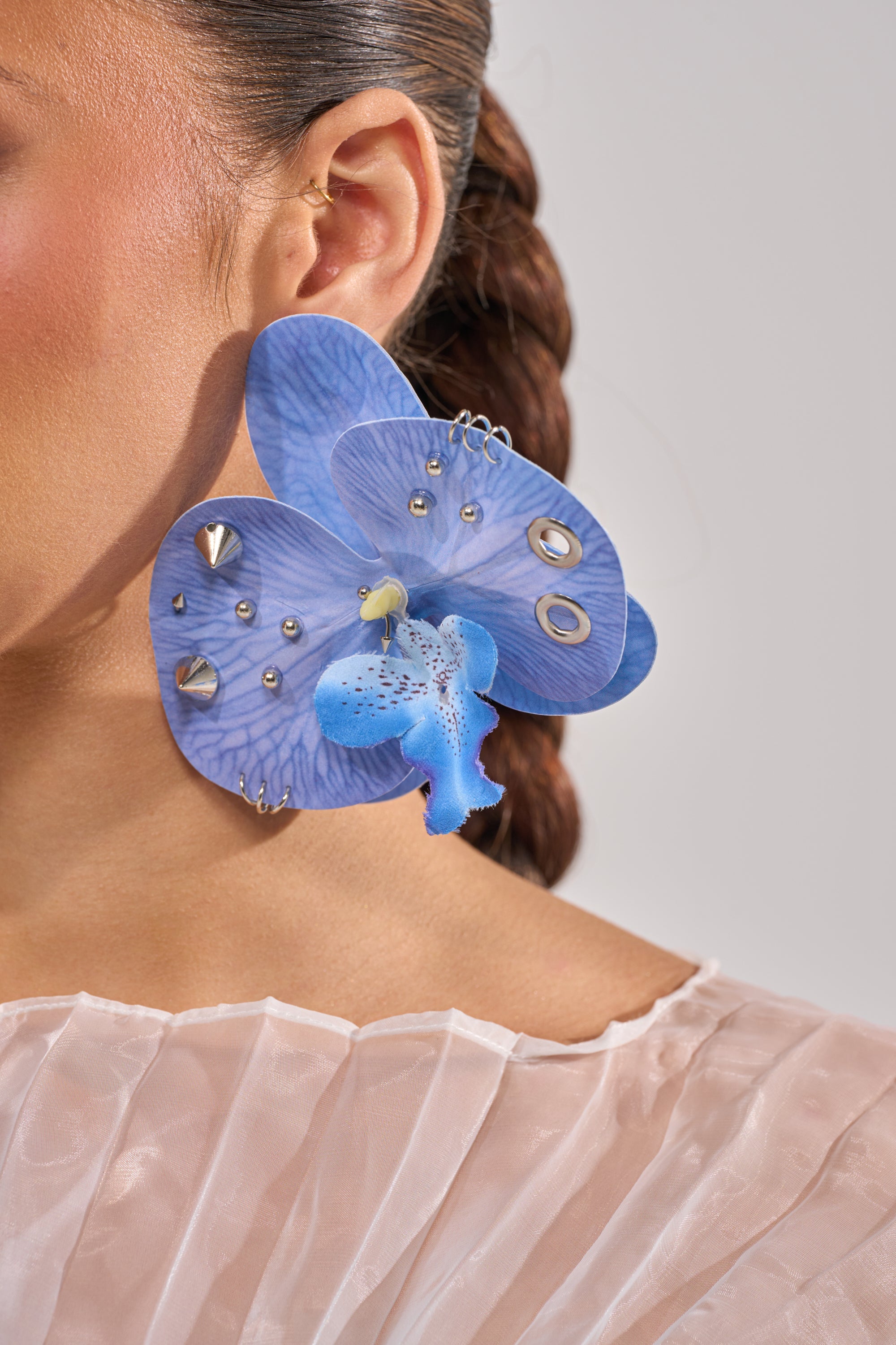 A woman wears the EDGY VIBES EARRING—a large, orchid-inspired statement piece in cobalt blue with metallic studs and hoop details. Her hair is pulled back, dressed in sheer pleated fabric, with only part of her face visible. 5