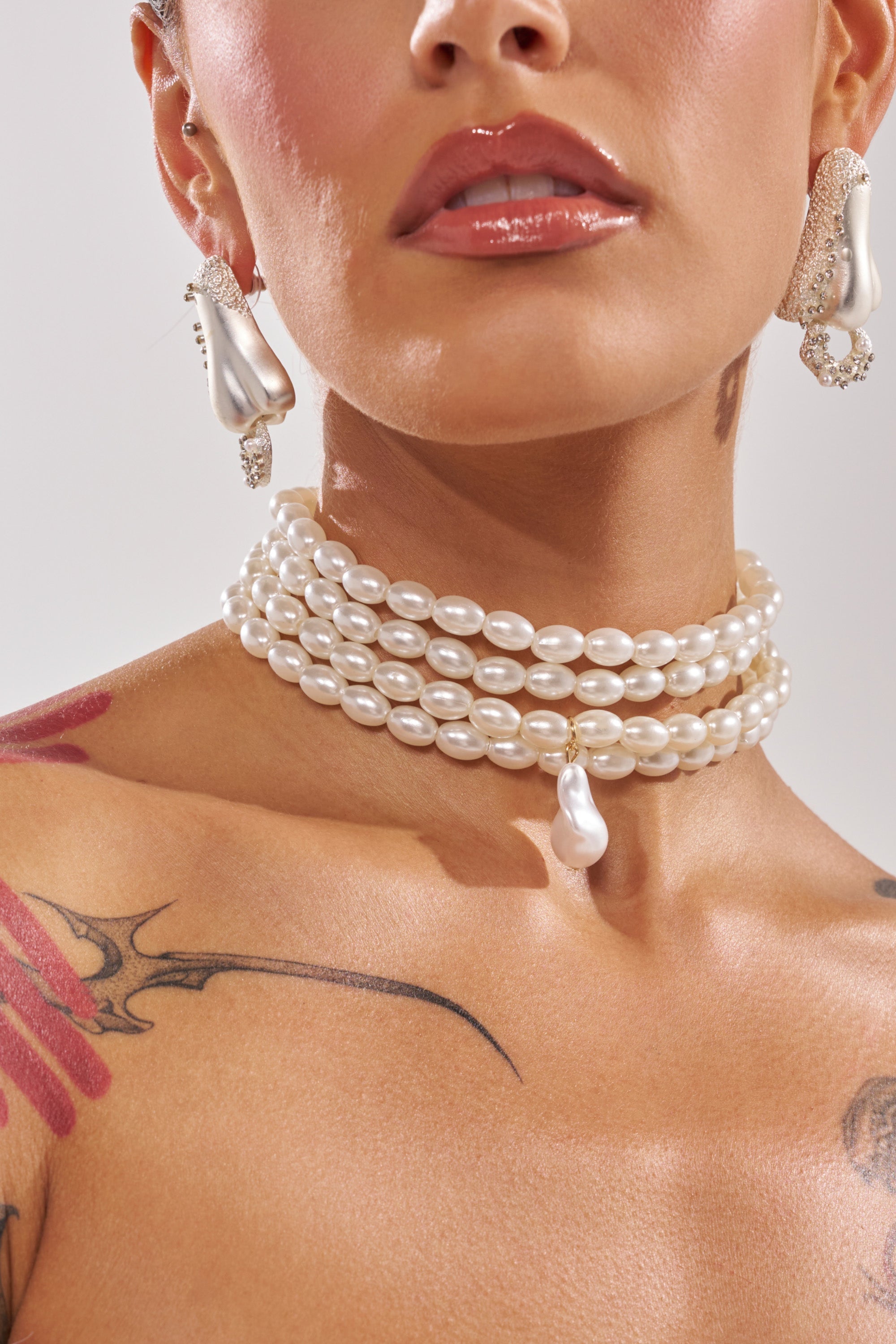 A woman flaunts tattoos on her shoulder and chest while wearing the IN THE OCEAN NECKLACE, a layered faux pearl choker with a teardrop pendant, matching pearl drop earrings, glossy lips, and stud ear piercings. 0