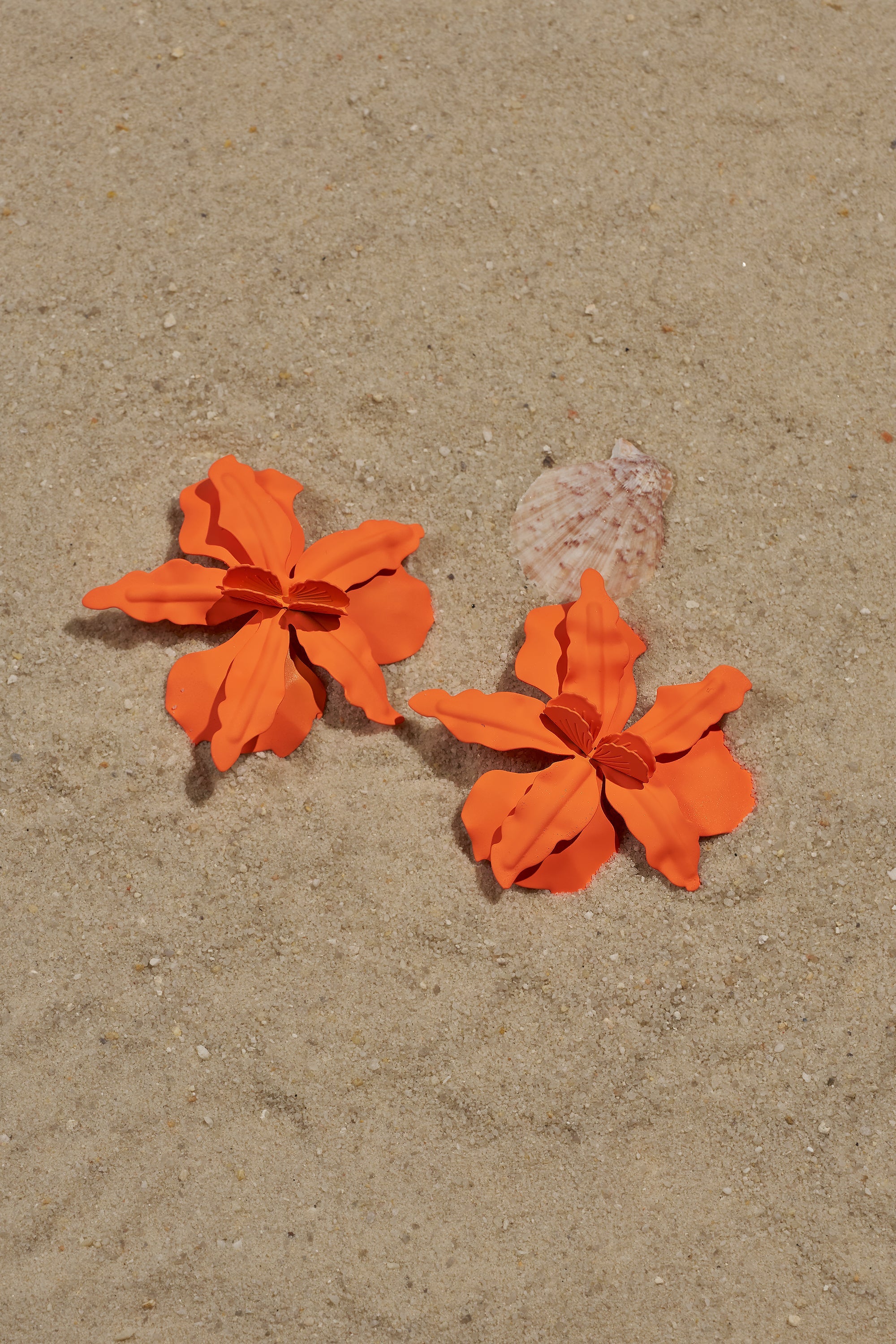The IDK U EARRING, in bright orange with a bold floral design, lies next to a seashell on smooth, light tan sand, showcasing the playful charm of oversized statement earrings. 0