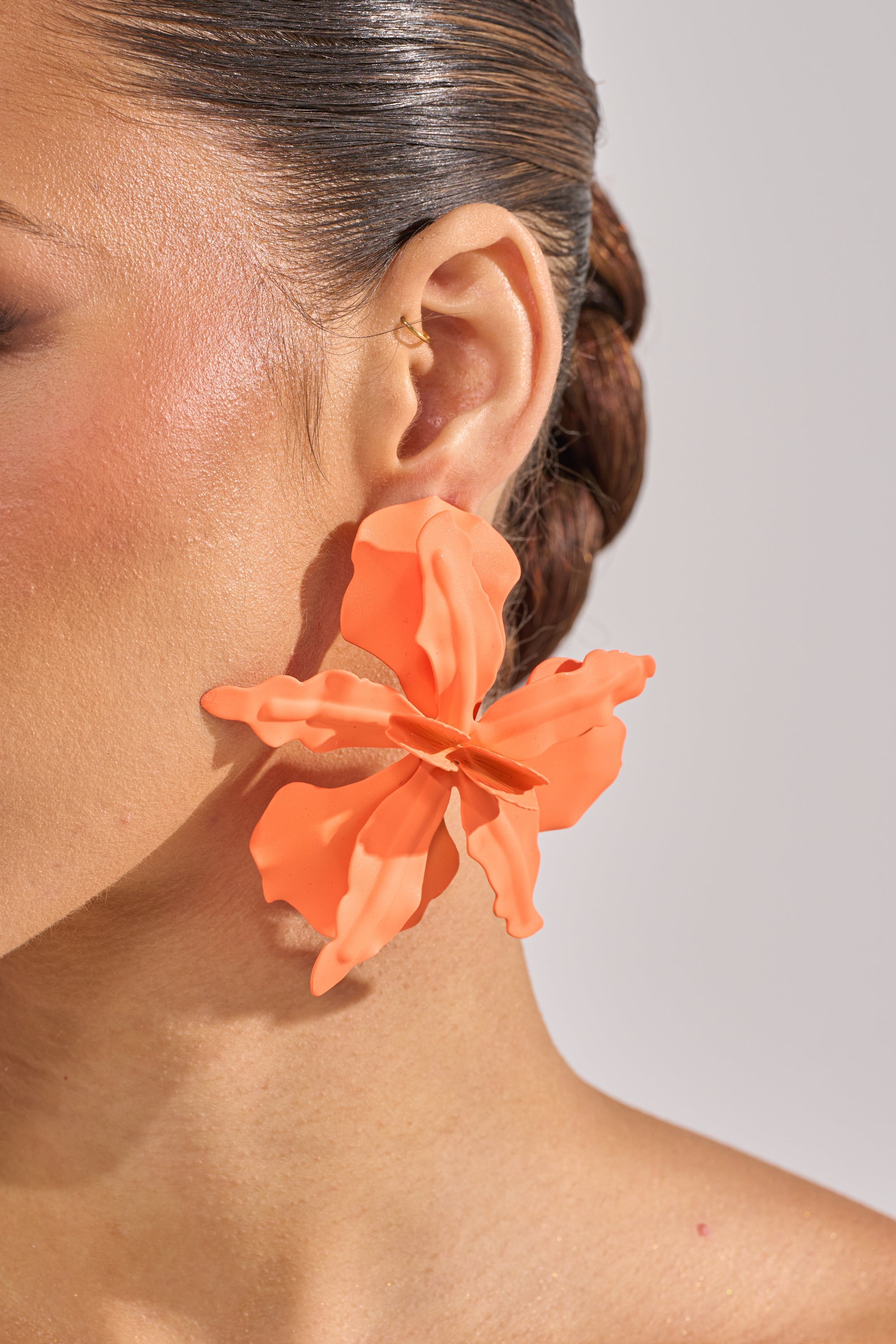 Close-up of a woman's ear wearing the IDK U EARRING—an oversized vibrant orange floral earring—with a small gold hoop above. Her sleek, pulled-back hair and naturally glowing skin highlight the statement piece. 3