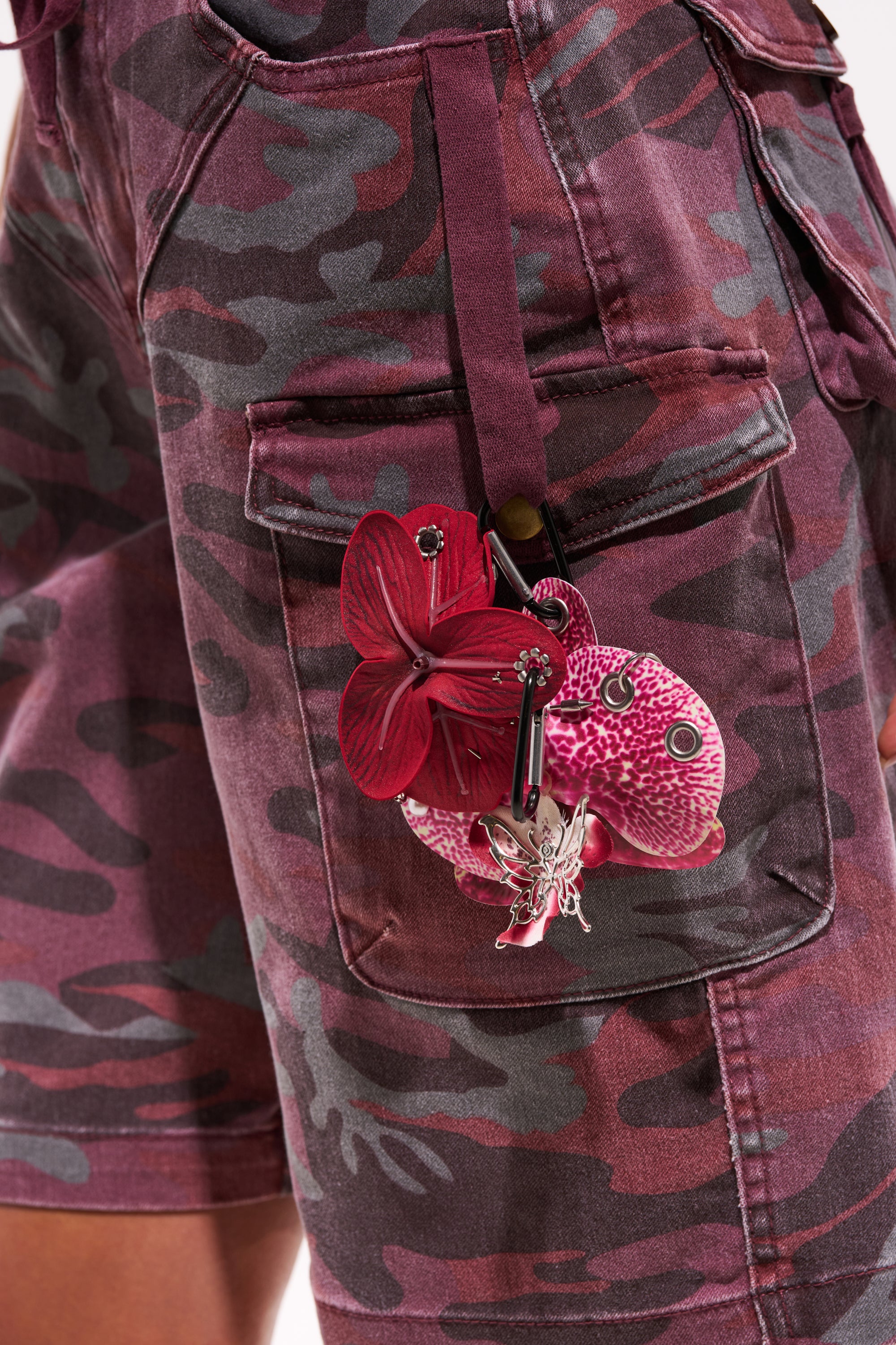Close-up of a person wearing purple and dark red camo shorts with a side pocket, featuring the PIERCED ORCHID FLOWER KEYCHAIN SET adorned with Y2K-inspired floral charms, including an orchid, pink-white mushroom, and silver charm. 0