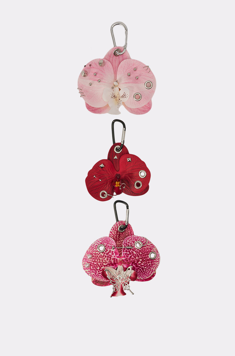 The PIERCED ORCHID FLOWER KEYCHAIN SET features three Y2K-inspired orchid keychains in pink, red, and purple with metallic studs or floral charms, displayed vertically on a plain white background. 4
